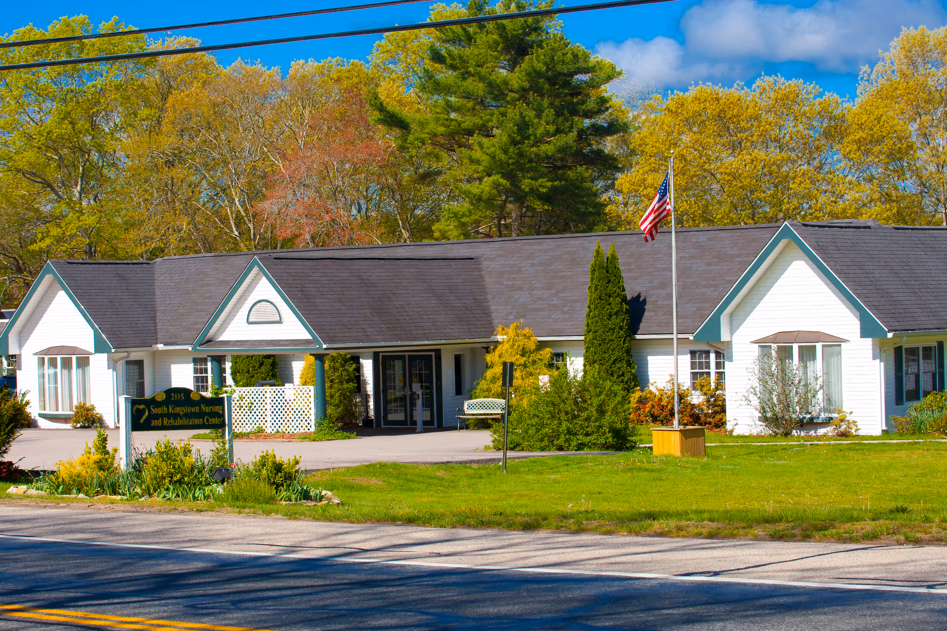 Front view of the South Kingstown Nursing & Rehabilitation Center, a single-story white building with an American flag and landscaped lawn.