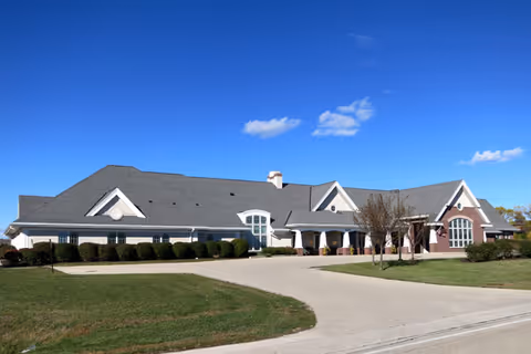 Front exterior of a single-story senior living facility with a curved driveway, manicured lawn, and clear blue sky.