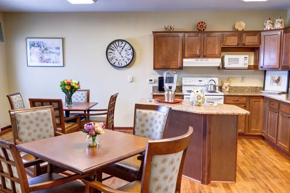 Bright communal dining area with multiple tables and chairs, floral centerpieces, and a kitchen island with wooden cabinets in the background.