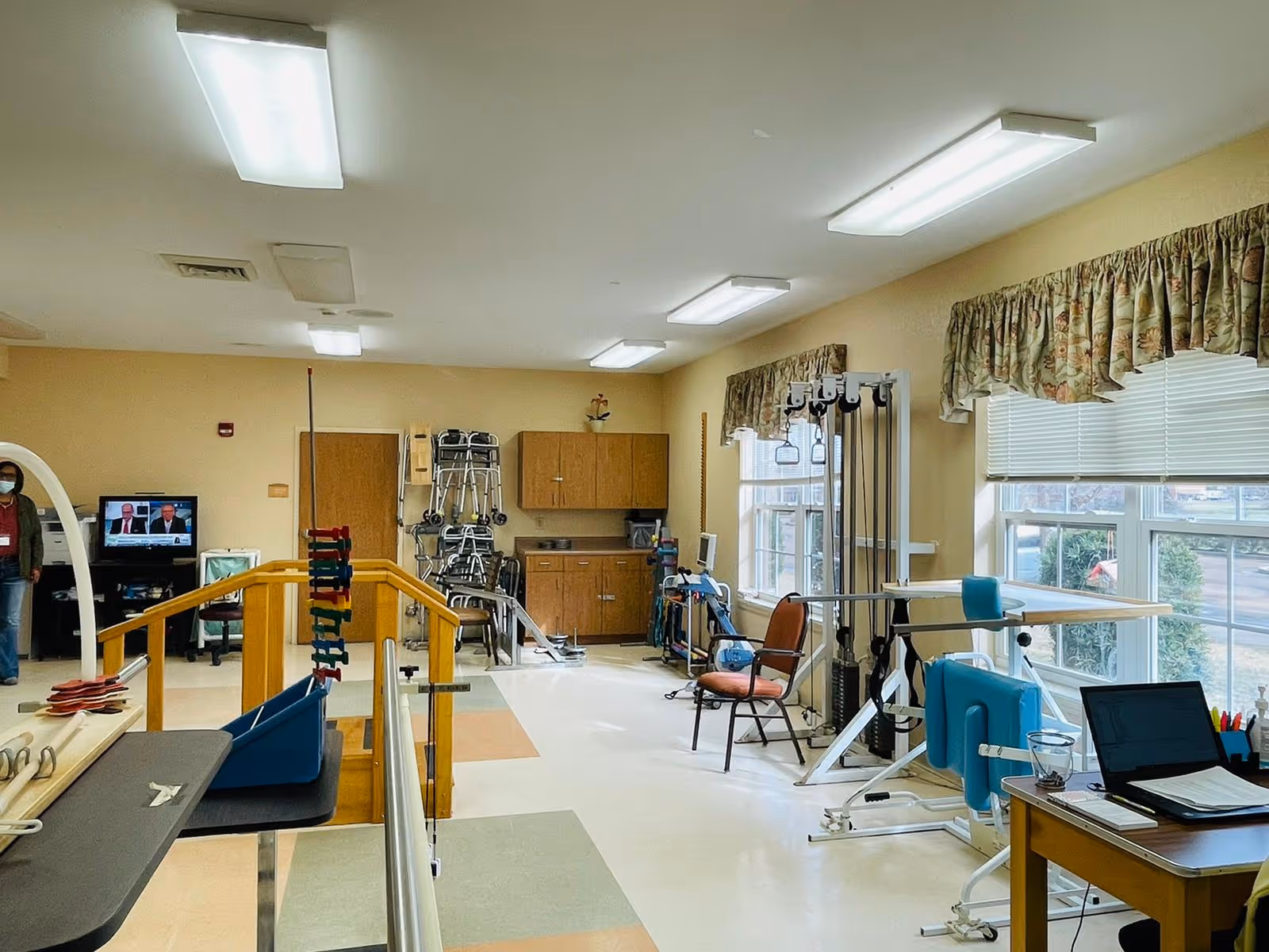 Interior view of a rehabilitation and healthcare center room with physical therapy equipment including parallel bars, exercise machines, chairs, and a desk with a laptop. There are large windows with floral curtains letting in natural light, and a person wearing a mask is visible near a television in the background.