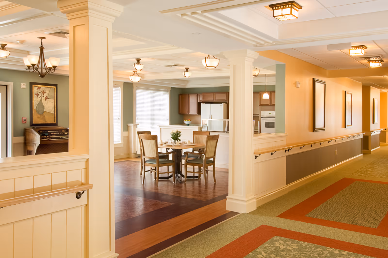 Interior view of a senior living facility showing a hallway with handrails and a dining area with a round table and four chairs. The dining area is adjacent to a kitchen with wooden cabinets and a white refrigerator. The space is well-lit with ceiling lights and has warm, neutral colors on the walls and carpet.