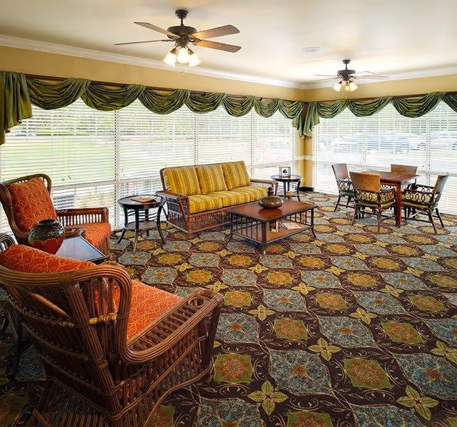 Sunlit lounge with wicker seating, a striped sofa, coffee tables and a dining table on a patterned carpet in front of large windows with green valances.