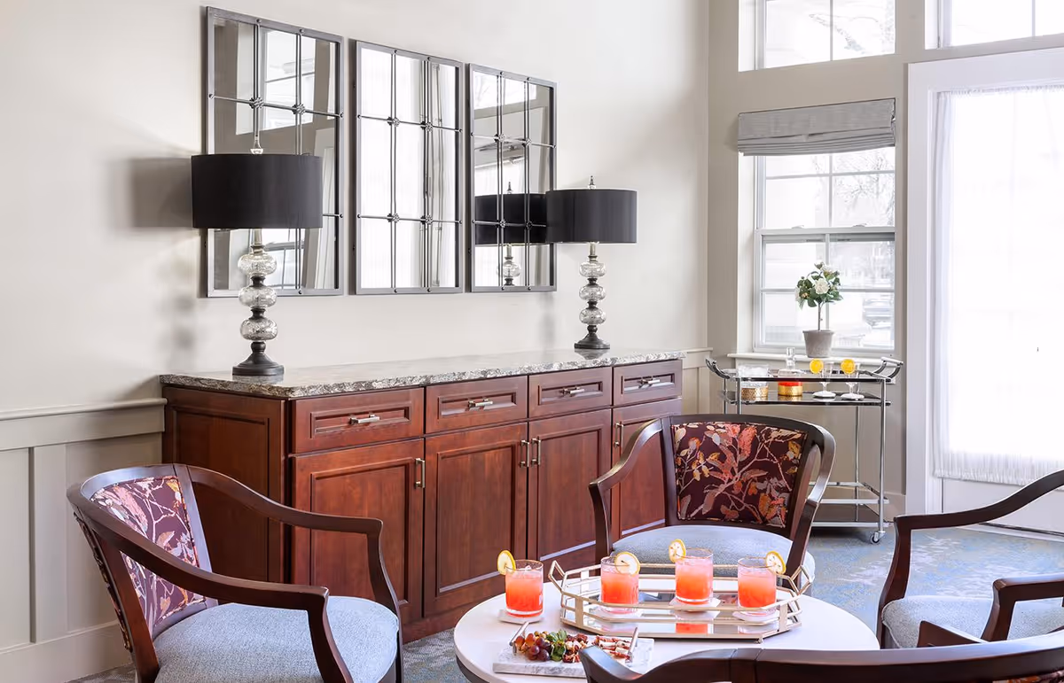 A bright sitting area with upholstered chairs around a coffee table, a wooden sideboard with lamps and mirrored wall panels, and a beverage cart by a window.
