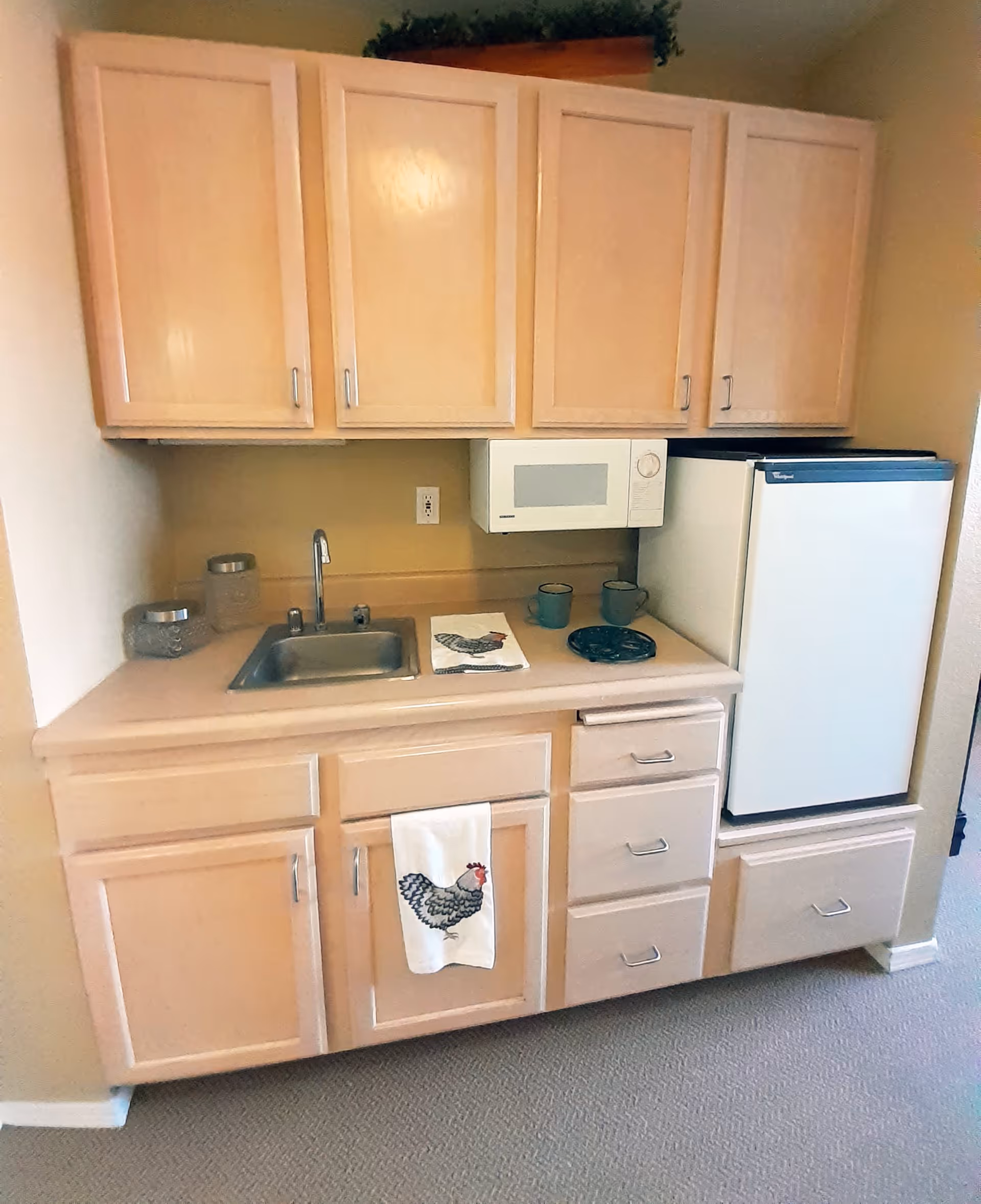 Small kitchenette with light wood cabinets, a stainless steel sink, a white microwave mounted above the counter, a compact white refrigerator, and a beige countertop with two mugs and a decorative towel featuring a chicken design.