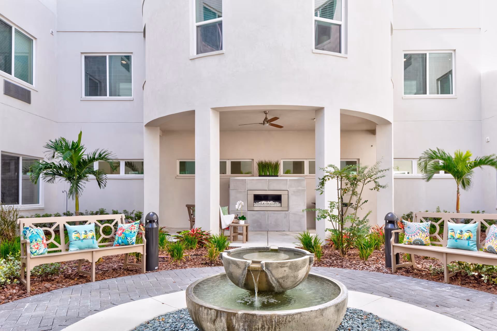 Outdoor courtyard area with a round two-tier stone water fountain in the center, surrounded by a paved walkway. There are two benches with colorful tropical-themed cushions on either side, green plants, and small palm trees. The background features a light-colored building with multiple windows and a covered patio area with a ceiling fan and a modern fireplace.