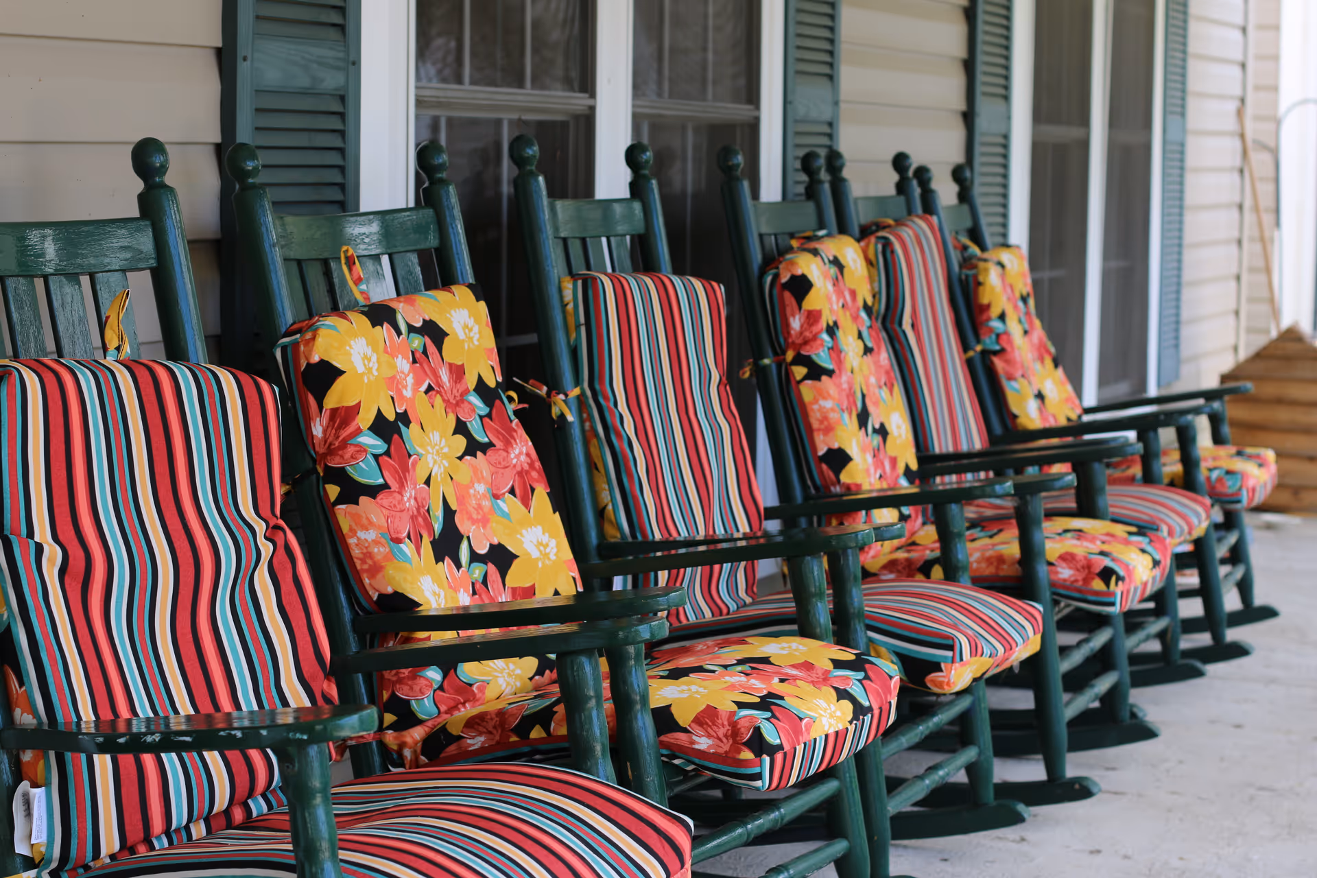 A row of green wooden rocking chairs with colorful striped and floral cushions lined up on a covered porch with beige siding and windows with green shutters.