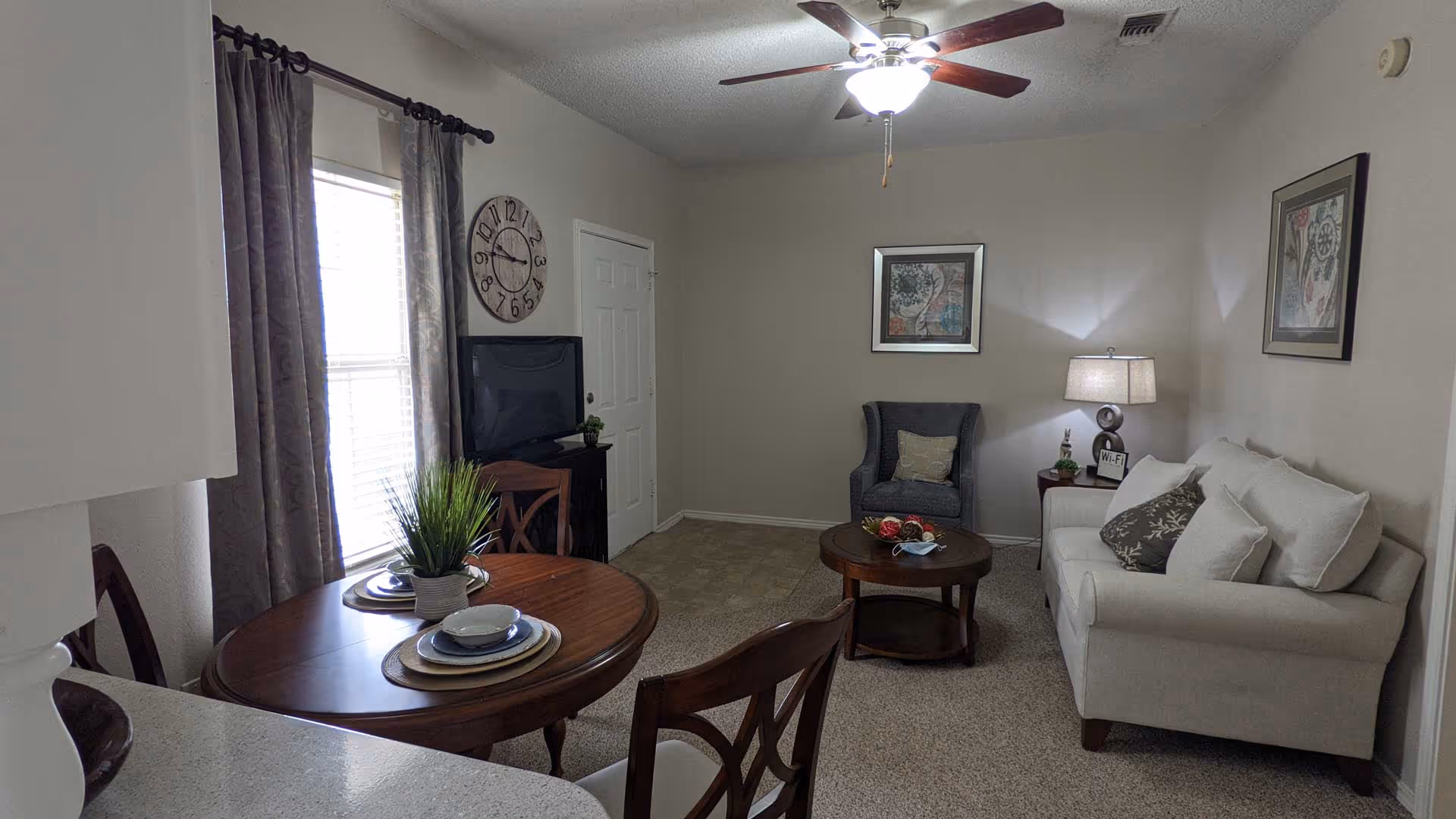 A cozy living room with a white sofa adorned with pillows, a dark armchair, and a round wooden coffee table with decorative items. A wooden dining table with place settings and a potted plant is near a window with curtains. A ceiling fan with lights is on the ceiling, and framed artwork hangs on the walls. A TV is placed on a stand near a door.