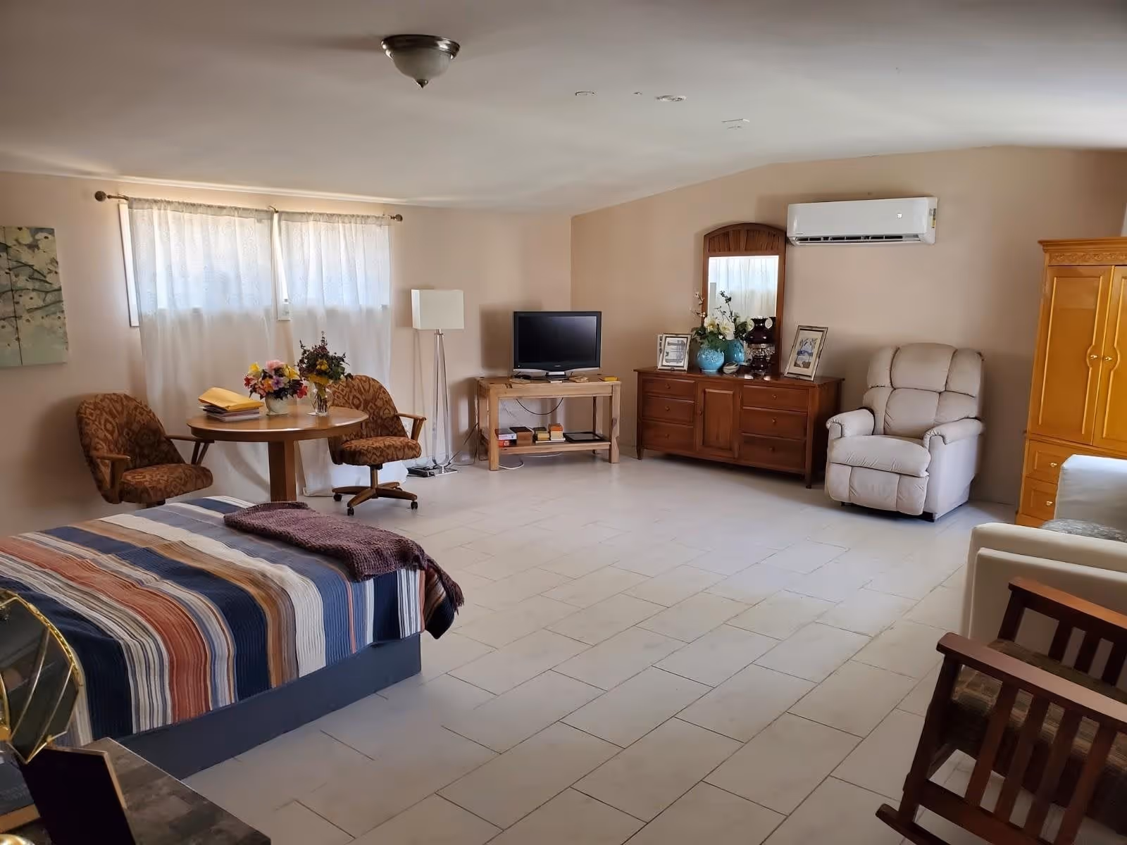 A spacious room with a bed covered in a striped blanket, a small round table with two patterned chairs, a floor lamp, a TV on a wooden stand, a wooden dresser with a mirror and decorative items, a beige recliner chair, and a wooden wardrobe. The room has light-colored tiled flooring and beige walls with a window covered by white curtains.