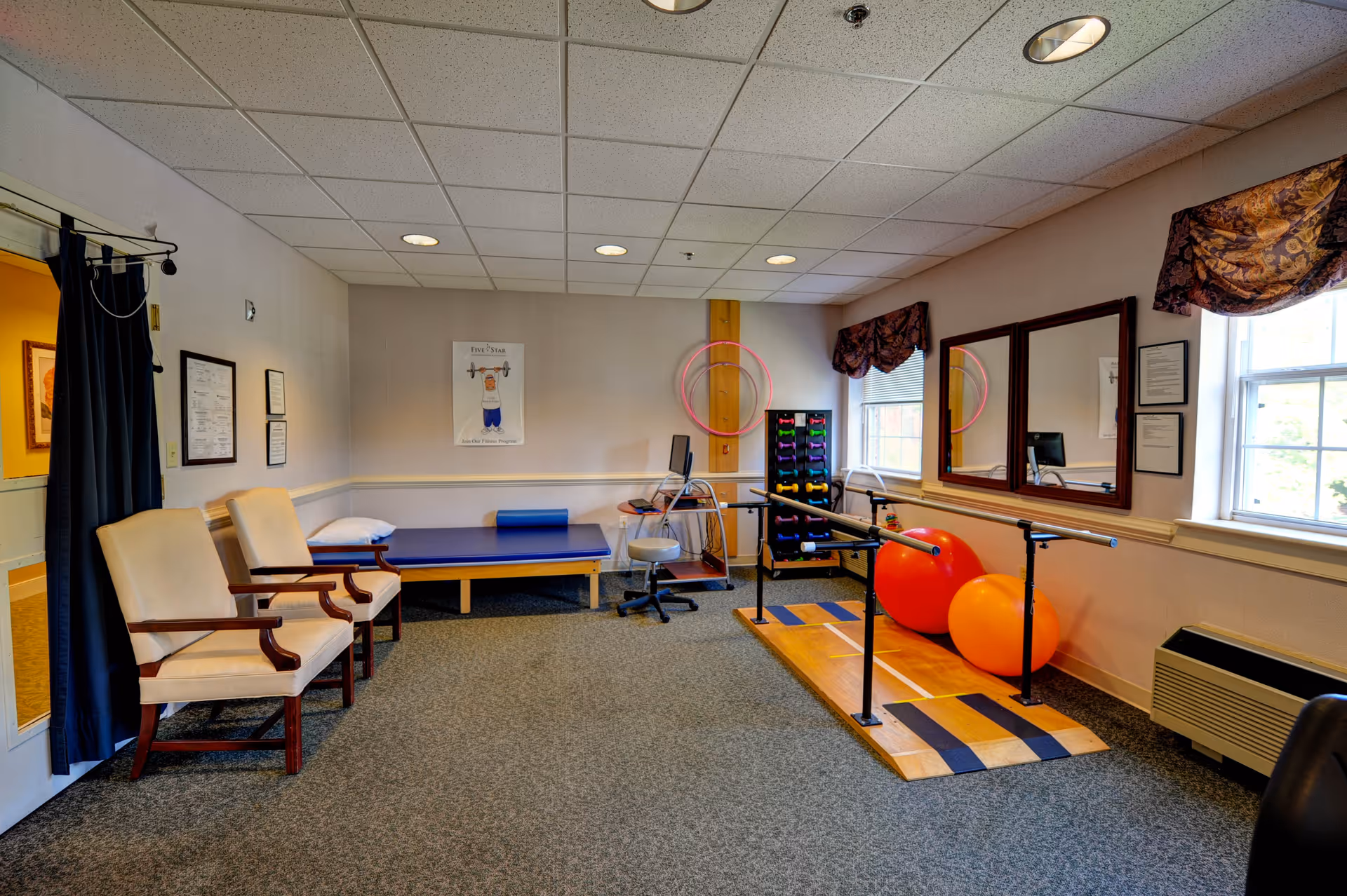 A physical therapy or exercise room with two beige armchairs on the left, a blue padded therapy table with a pillow, a computer workstation, a rack of colorful dumbbells, two large exercise balls, and parallel bars on a wooden platform. The room has carpeted floors, beige walls with a chair rail, two windows with patterned valances, and a large mirror on the right wall.