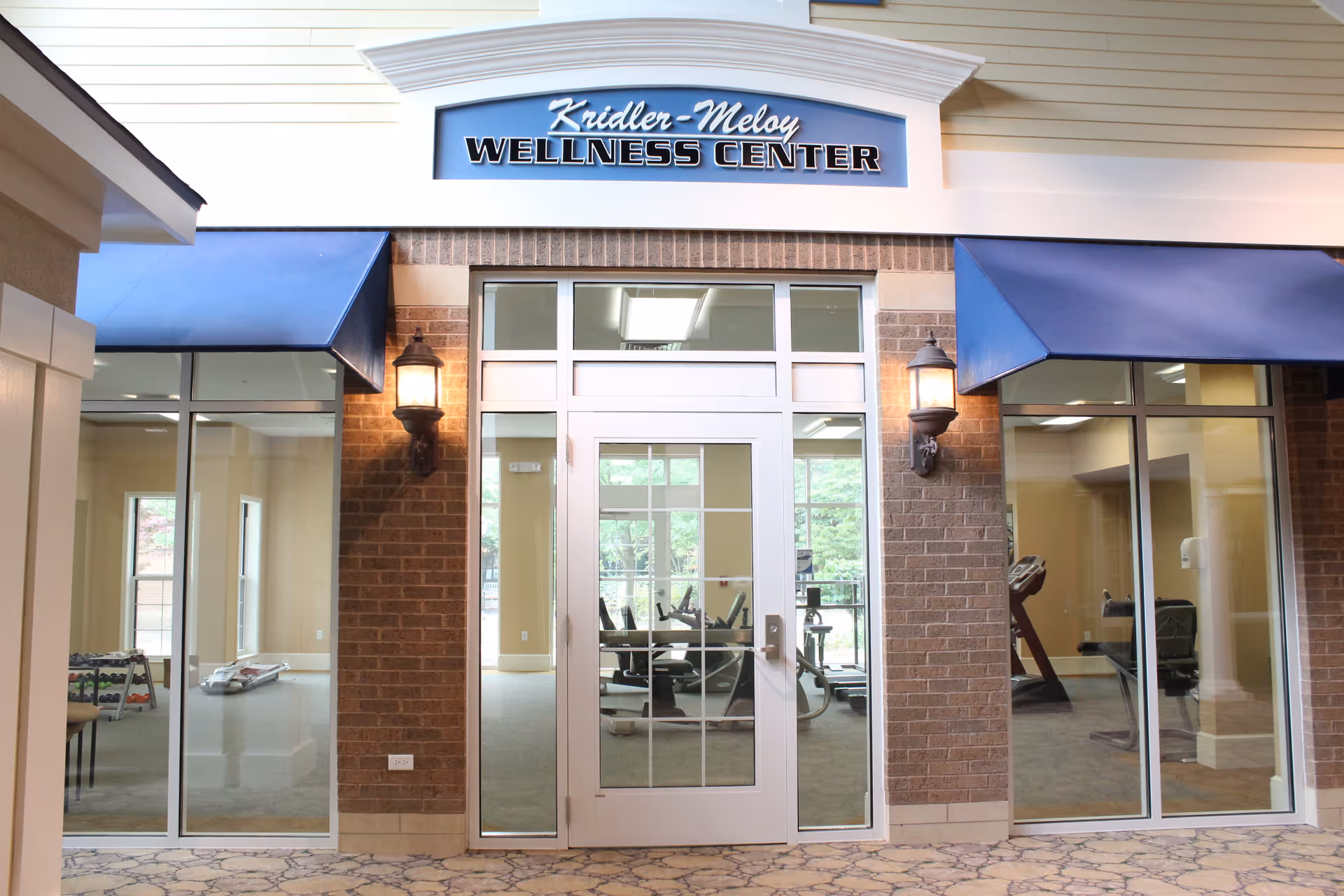 Entrance to the Kridler-Meloy Wellness Center at Ohio Living Swan Creek, featuring a glass door and windows with exercise equipment visible inside. The exterior wall is brick with two lit wall-mounted lanterns and blue awnings above the windows.