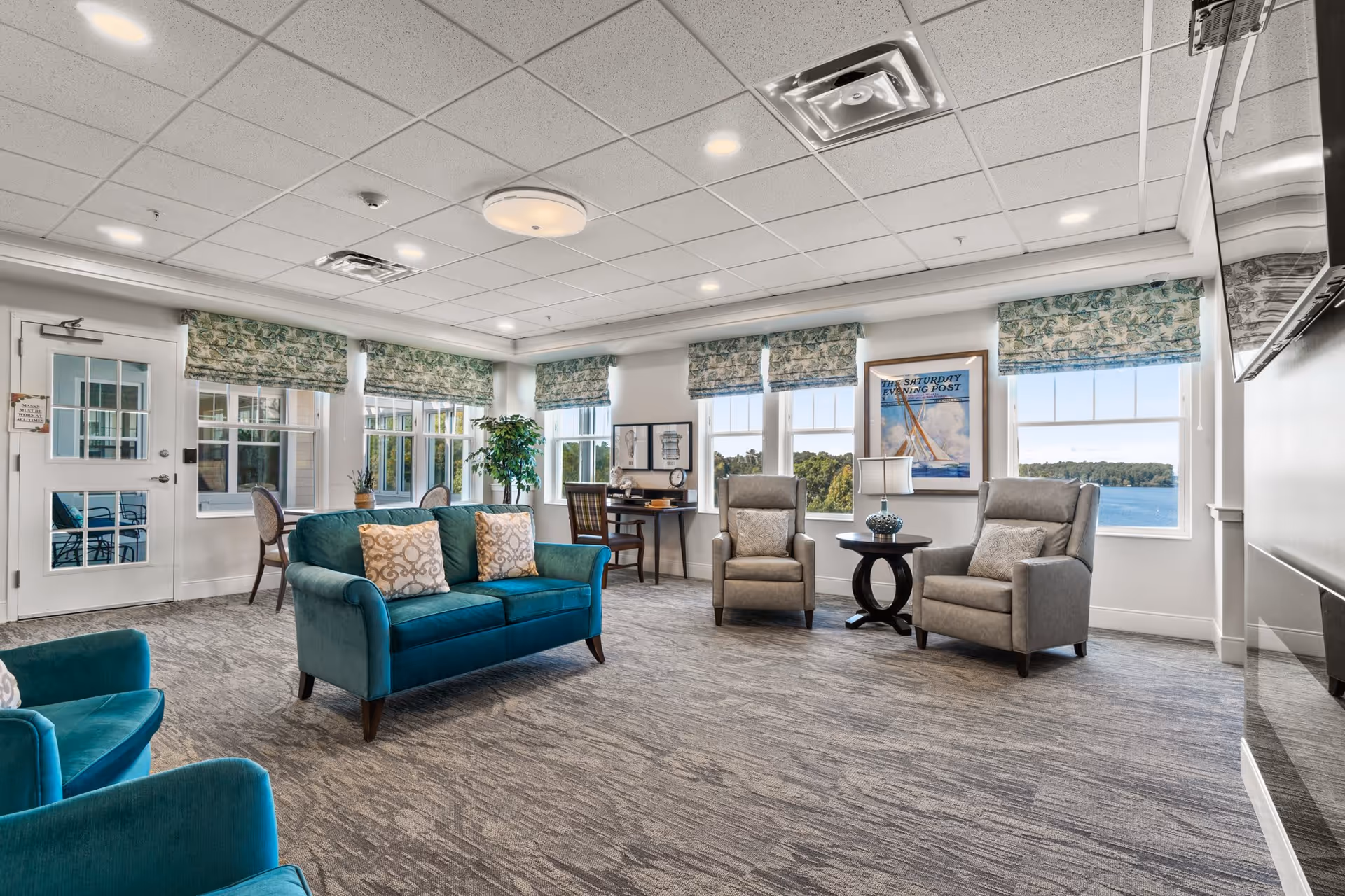 A bright and spacious senior living common area with large windows offering a view of a lake. The room features teal upholstered sofas and armchairs, two gray armchairs around a small round table with a lamp, a desk with a chair, and patterned window valances. The ceiling has recessed lighting and a central light fixture.