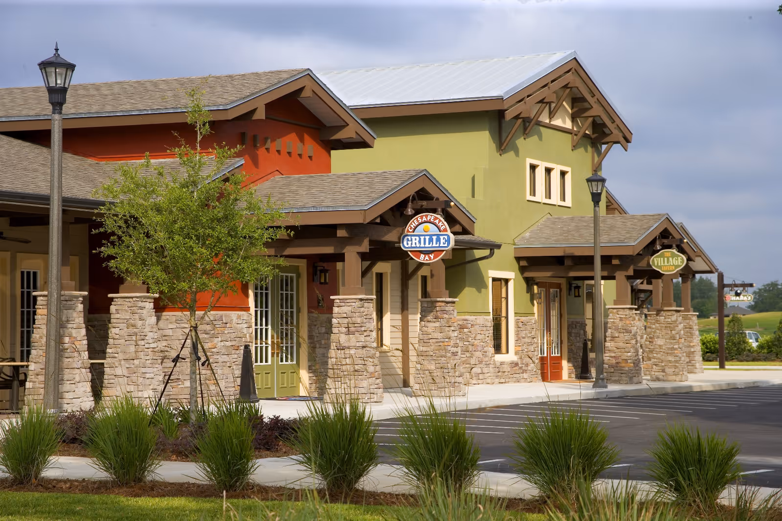 Exterior view of a senior living facility building with stone pillars and colorful walls in green and orange. There are signs for 'Chesapeake Bay Grille' and 'The Village' hanging from the building. The area includes a parking lot, lamp posts, and landscaped greenery with small trees and bushes.
