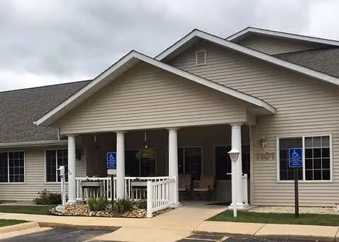 Exterior view of Arlington Place at Oelwein, showing the front entrance with a covered porch supported by white columns, two handicap parking signs, and a beige building with multiple windows under a cloudy sky.
