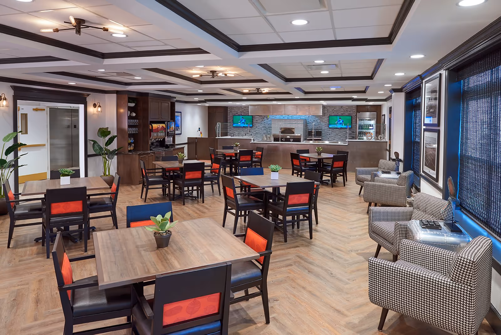 A spacious dining area with several wooden tables and black chairs with red cushions. The room features a modern ceiling with recessed lighting and dark wood trim. There are plants placed on some tables and near the walls. On the right side, there are comfortable patterned armchairs with small metallic side tables. The back wall has a serving counter with two televisions mounted above it and a beverage station to the left.