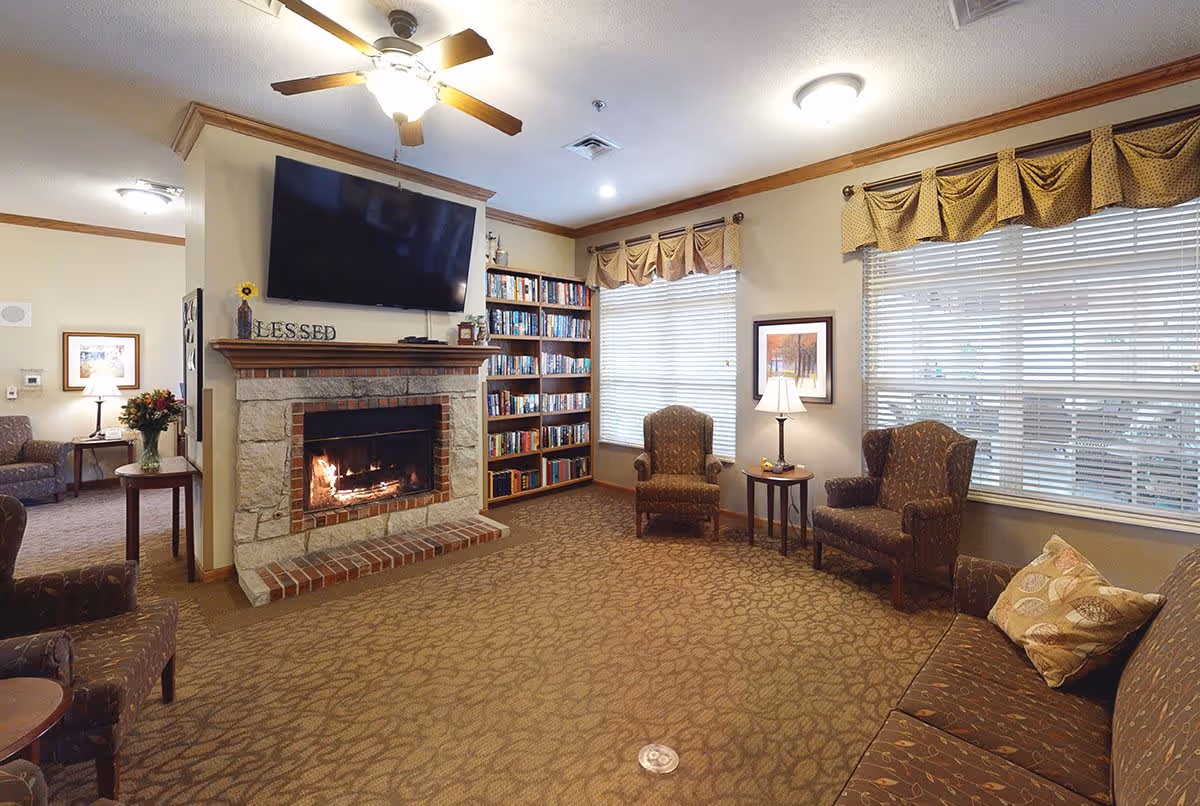 A cozy living room area in Trail Creek Place Memory Care featuring a stone and brick fireplace with a fire burning, a large flat-screen TV mounted above it, a bookshelf filled with books, several upholstered armchairs and a sofa with patterned cushions, two windows with blinds and valances, and warm lighting from ceiling fixtures and table lamps.