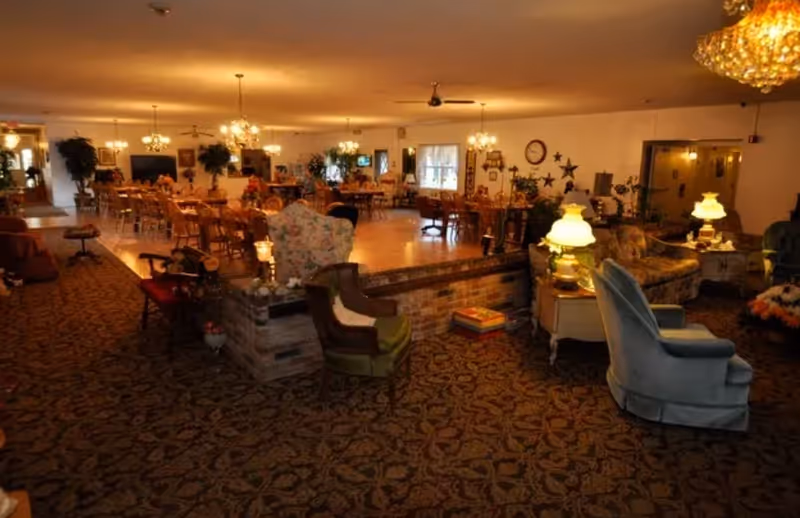 Spacious senior living common area with sofas, tables, lamps, plants, and warm lighting.