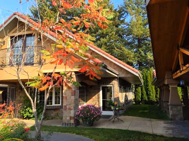 Exterior view of a residential-style building with a stone and brick facade, a red-tiled roof, and a small patio area with a table and potted flowers. There is a tree with red and green leaves in the foreground and tall green trees in the background under a clear blue sky.