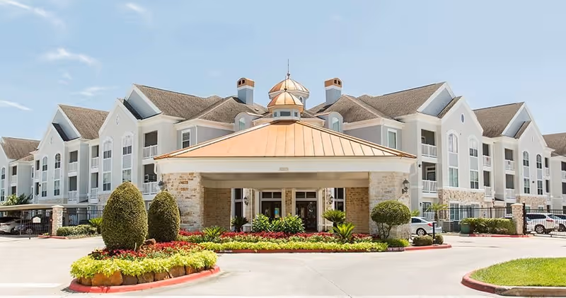 Front exterior view of The Grand Parkway senior living facility featuring a large entrance with a copper-colored roof, manicured landscaping with bushes and flowers, and a multi-story building with white walls and multiple windows in the background under a clear blue sky.