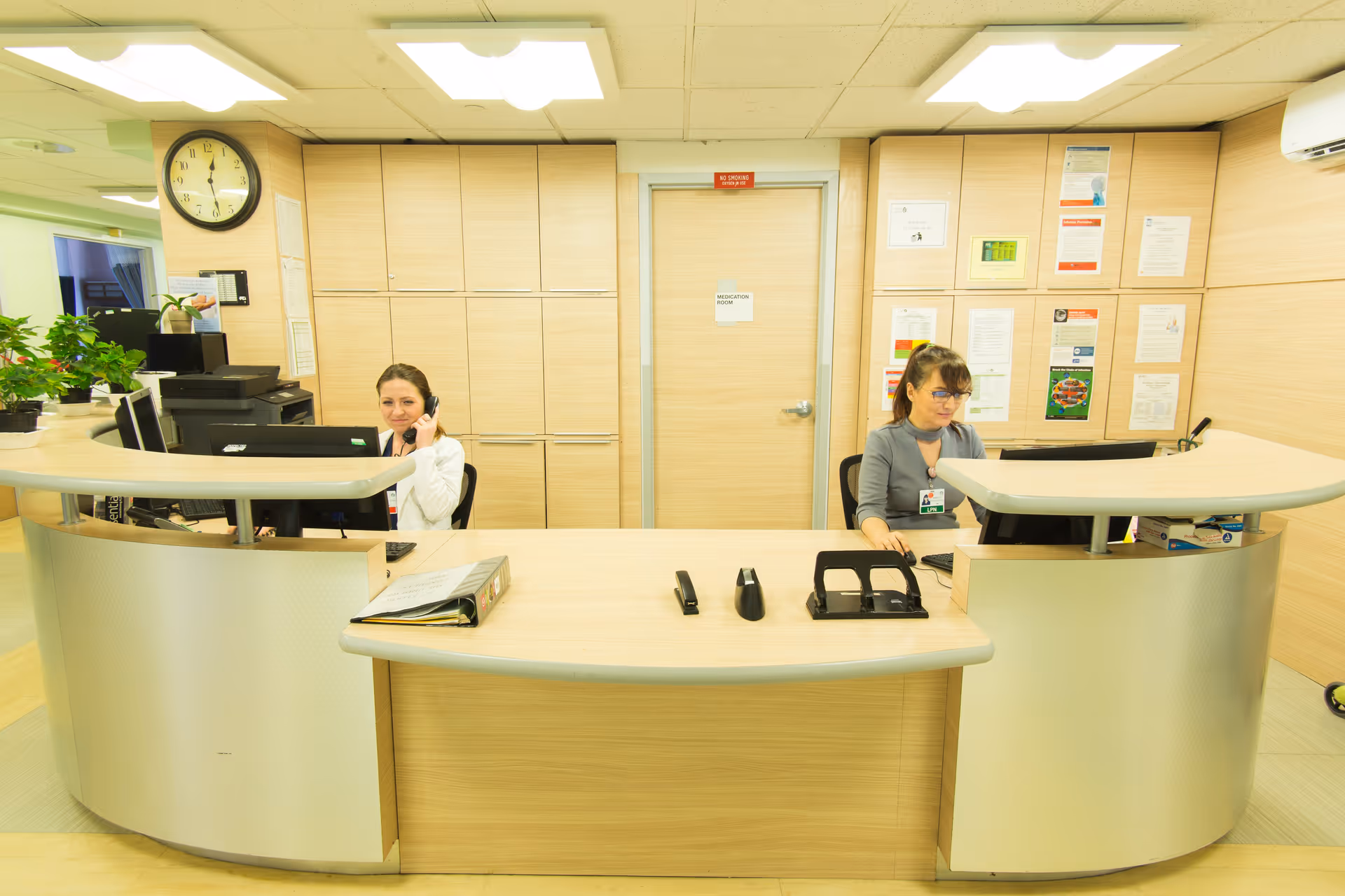Two reception staff seated behind a curved front desk in a bright nursing facility reception area.