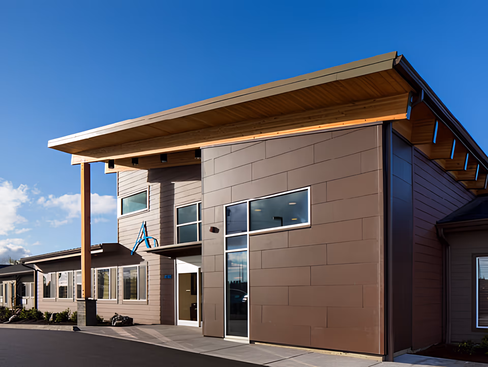 Front entrance of a modern building with a large overhanging roof, tall wooden support beam, and several windows.