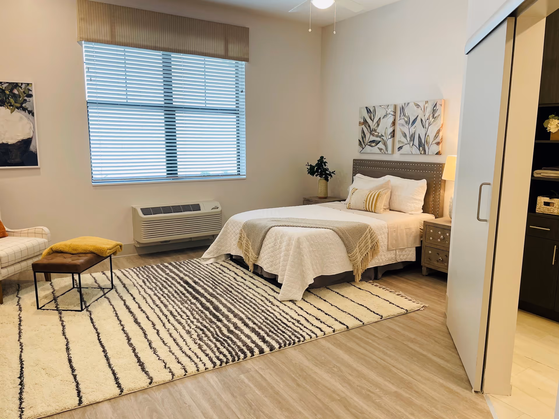 A cozy bedroom with a neatly made bed featuring white bedding and a beige throw blanket. There are two framed leaf-themed artworks above the bed, a nightstand with a lamp and a plant, a large window with blinds, a patterned area rug, a cushioned bench with a yellow throw, and a white armchair. The room has light wood flooring and a neutral color palette.
