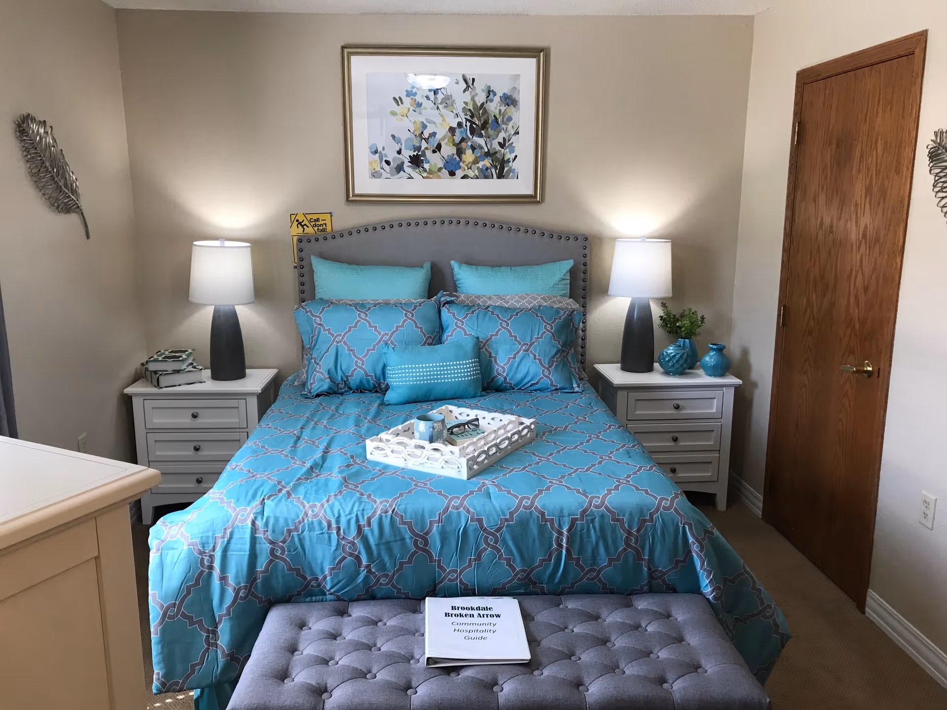 A neatly made bed with a turquoise and gray patterned comforter and pillows in a bedroom. Two white nightstands with lamps and decorative items flank the bed. A framed floral artwork hangs above the bed. A gray tufted bench is at the foot of the bed with a booklet titled 'Brookdale Broken Arrow Community Hospitality Guide' on it. The room has beige walls and a wooden door on the right.