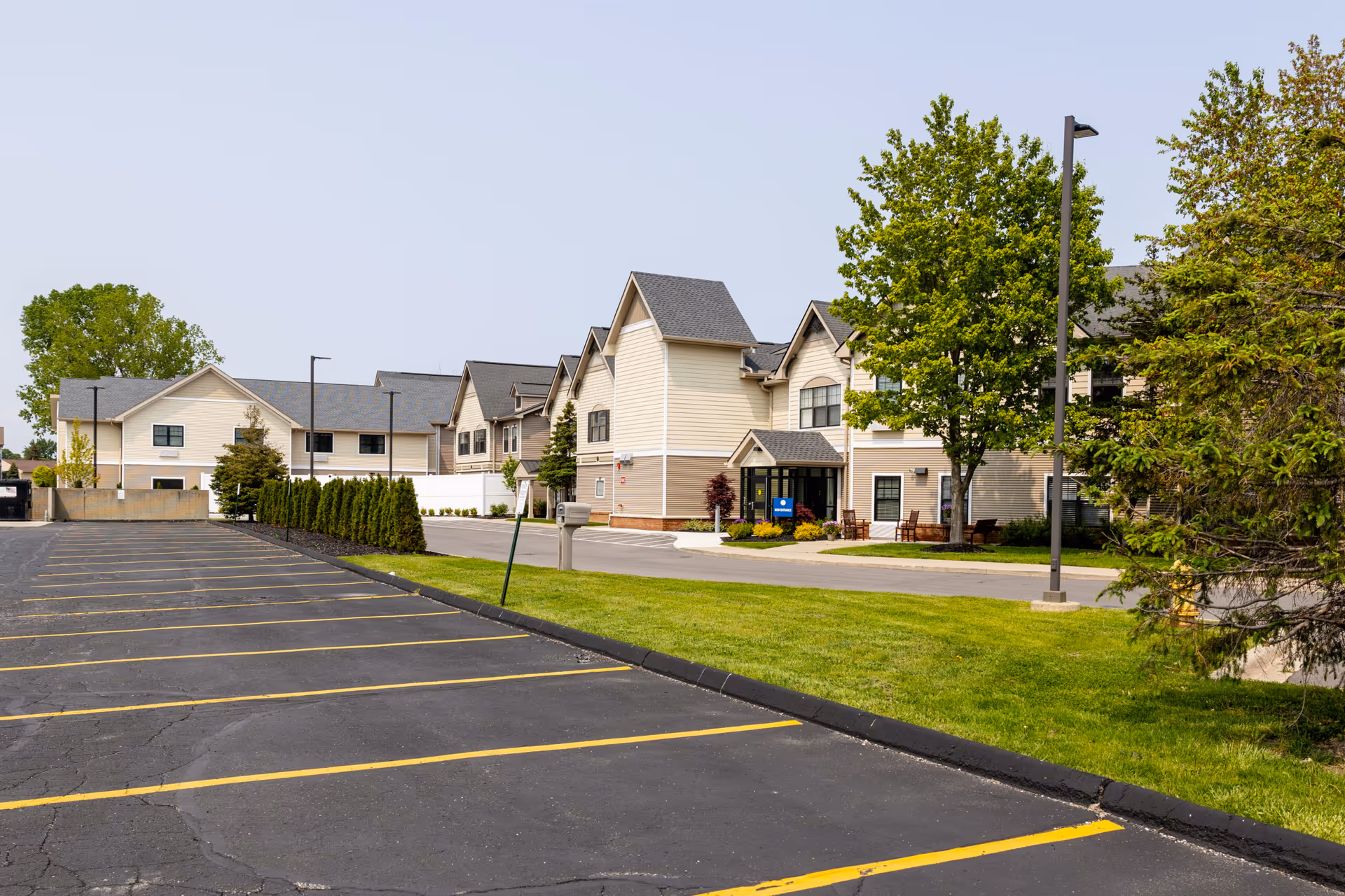 Exterior view of Commonwealth Senior Living at New Baltimore showing a large parking lot with yellow lines, a green lawn, trees, and a two-story beige building with multiple peaked roofs and windows under a clear sky.