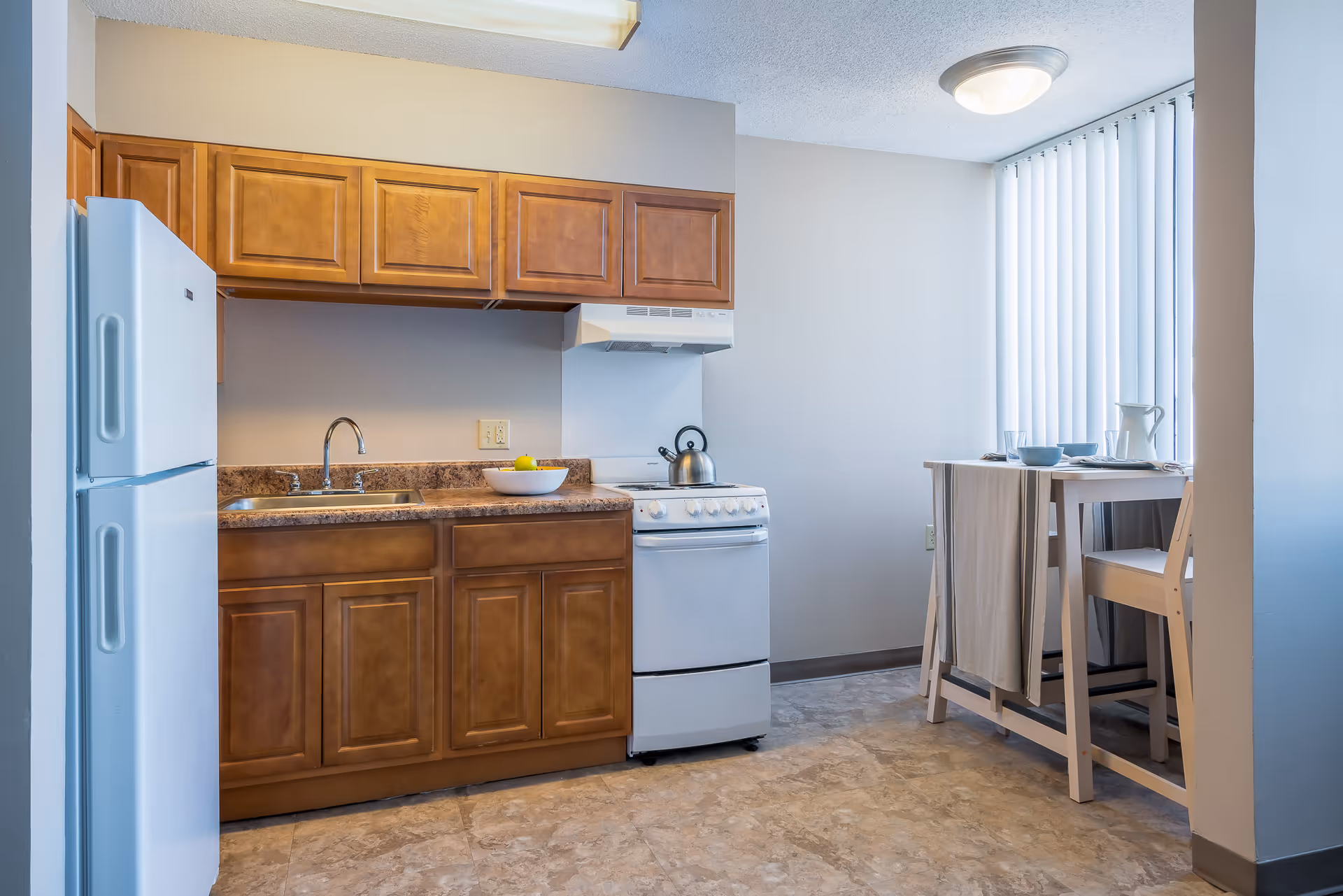Small kitchenette with wooden cabinets, a white refrigerator and stove, sink with countertop, and a small dining table by a window.