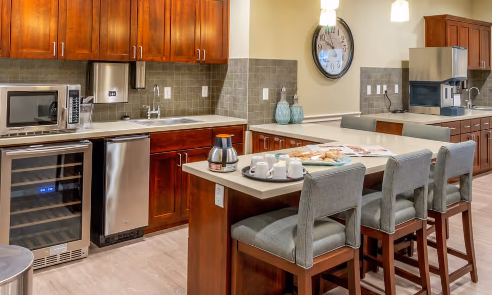 Communal kitchen area with an island and four upholstered barstools, stainless appliances, wooden cabinets, and a wall clock.