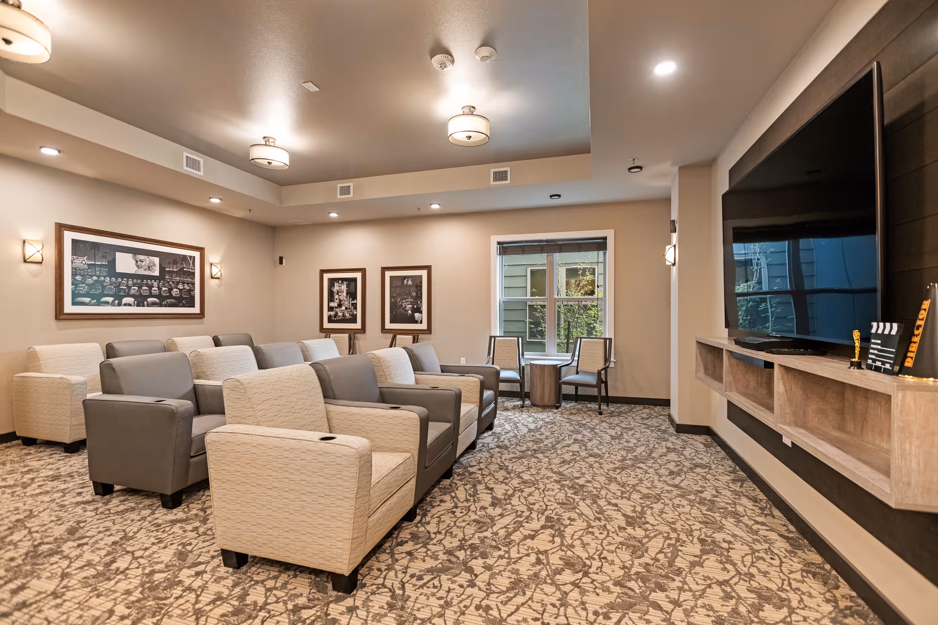 A cozy media room with multiple rows of cushioned armchairs facing a large flat-screen TV mounted on the wall. The room has patterned carpet flooring, beige walls adorned with framed black and white photographs, and ceiling lights providing warm illumination. There are two chairs and a small table near a window at the back of the room.