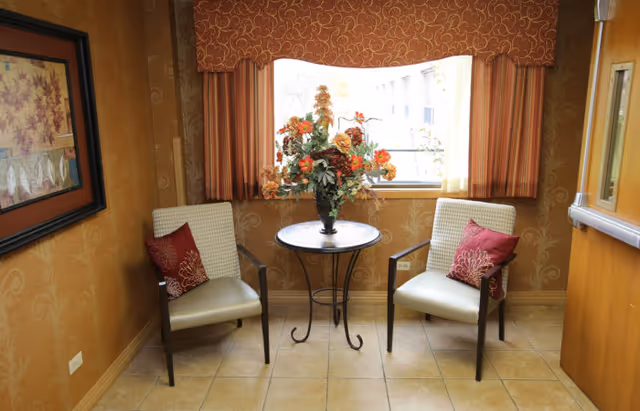 A small seating area with two beige armchairs featuring red decorative pillows, positioned on either side of a round metal table with a floral arrangement. The area has tiled flooring, patterned wallpaper, a window with red and gold curtains, and a framed artwork on the left wall.