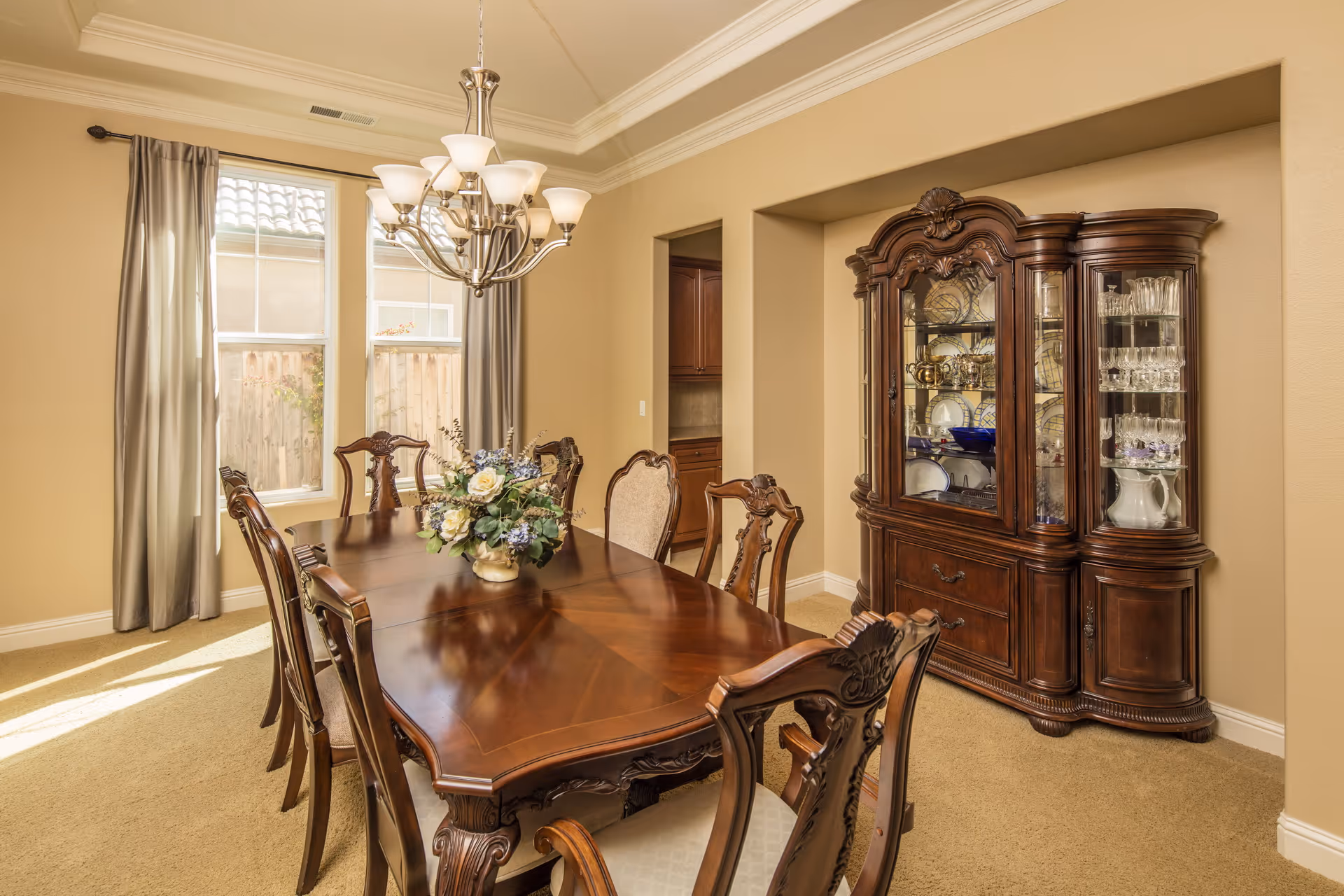 A well-lit dining room with a large wooden dining table surrounded by eight ornate chairs. A floral centerpiece decorates the table. Behind the table, there is a large wooden china cabinet filled with glassware and dishes. Two tall windows with gray curtains allow natural light to enter the room. A chandelier with multiple lights hangs above the dining table. The walls are painted beige and the floor is carpeted.
