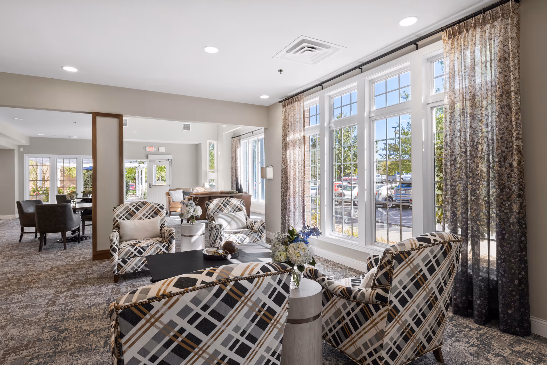 A bright and spacious senior living facility common area with large windows letting in natural light. The room features patterned armchairs arranged around a black coffee table with a floral centerpiece. Sheer curtains frame the windows, and additional seating areas with tables and chairs are visible in the background.