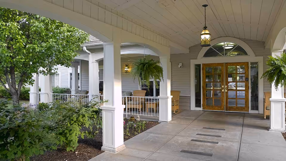 Covered entrance area of a senior living facility with white columns, hanging ferns, wicker chairs, and double wooden doors with glass panels. There are green bushes and a tree on the left side of the walkway leading to the entrance.