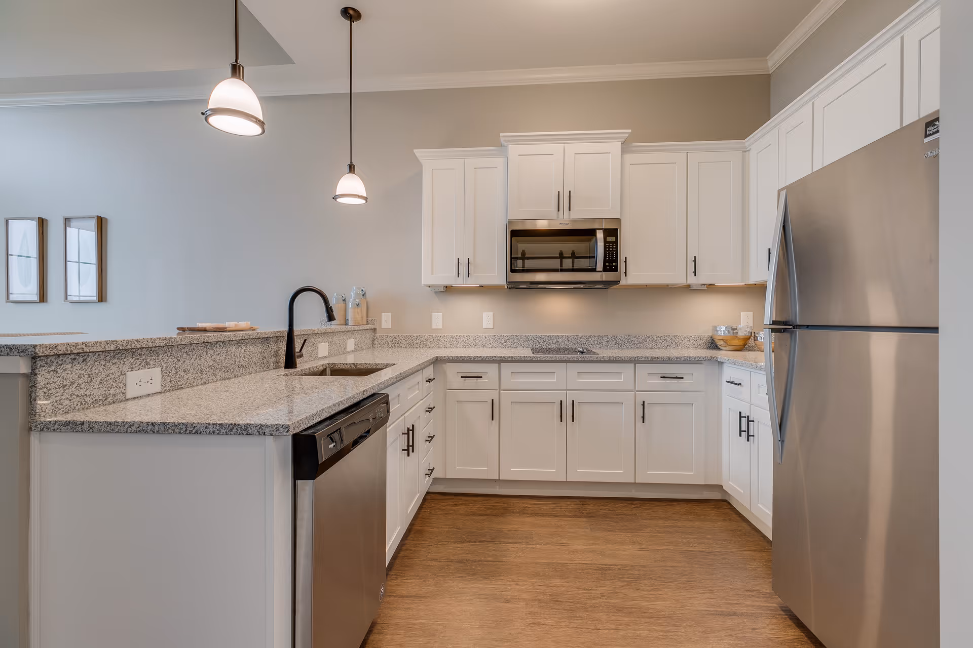 Modern kitchen with white cabinets, granite countertops, stainless steel refrigerator, dishwasher, and microwave. Two pendant lights hang over the counter, and there are two framed pictures on the wall in the background.