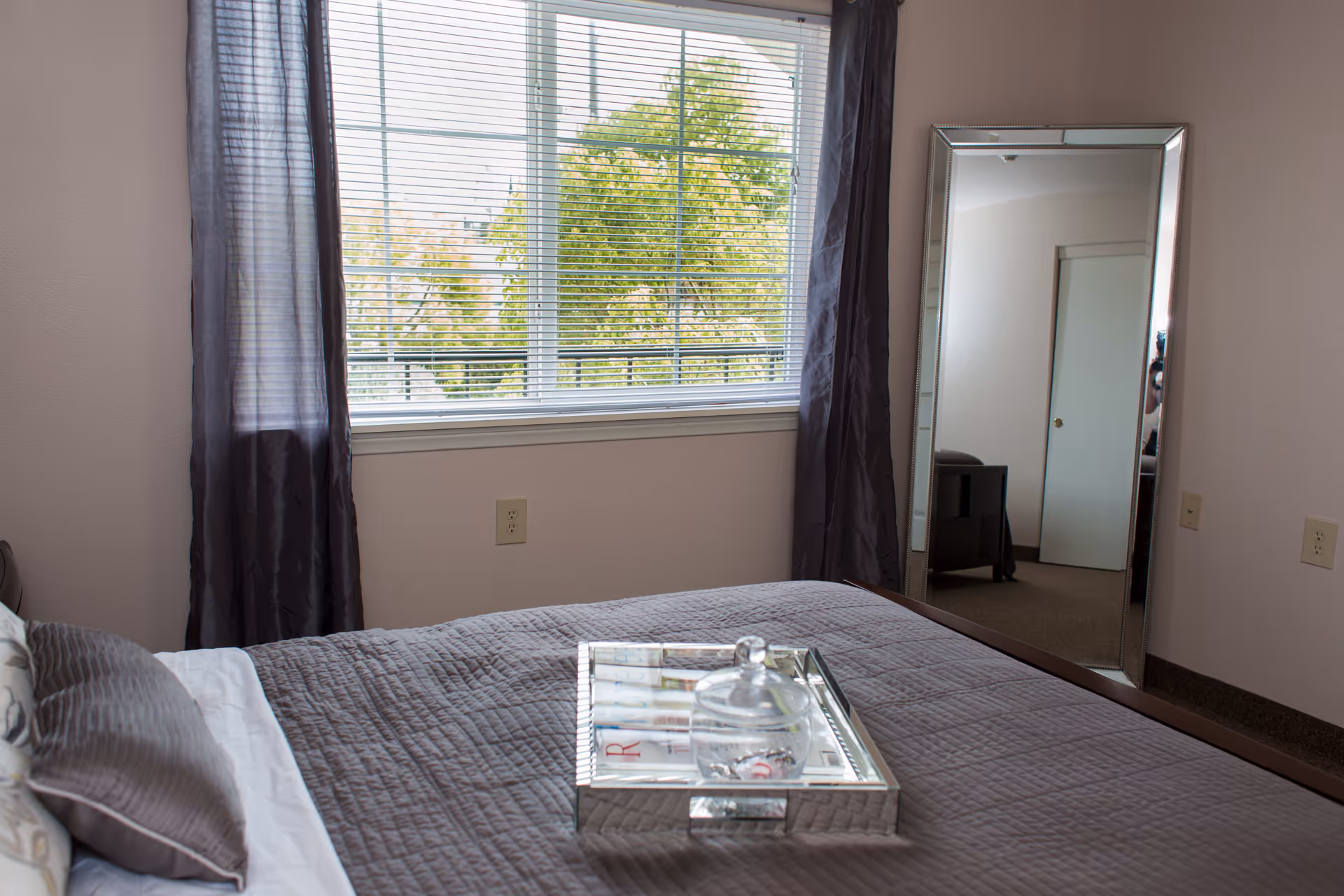 A bedroom with a neatly made bed covered in a gray quilt and pillows. A silver tray with a glass jar is placed on the bed. There is a large window with blinds and dark curtains, and a tall mirror leaning against the wall reflecting part of the room including a door and a small piece of furniture.
