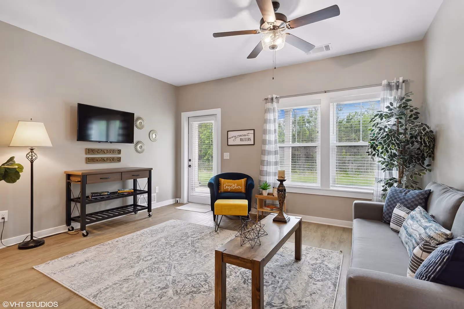 A bright and cozy living room with a gray sofa adorned with multiple cushions, a wooden coffee table with decorative items, a blue and yellow armchair, a floor lamp, a wall-mounted flat screen TV, and large windows with white blinds and sheer curtains letting in natural light. A ceiling fan with a light fixture is mounted on the white ceiling, and a door with glass panels leads outside.