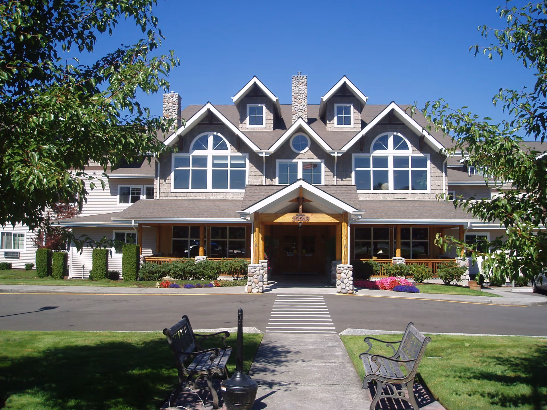 Front exterior view of a large, multi-story senior living facility building with stone chimneys, large windows, and a covered entrance. There are two benches on either side of a walkway leading to the entrance, surrounded by green grass, trees, and colorful flower beds under a clear blue sky.