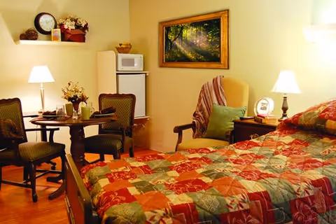 A cozy senior living bedroom with a quilted bed, armchair, small dining table and chairs, and a mini-fridge with a microwave.