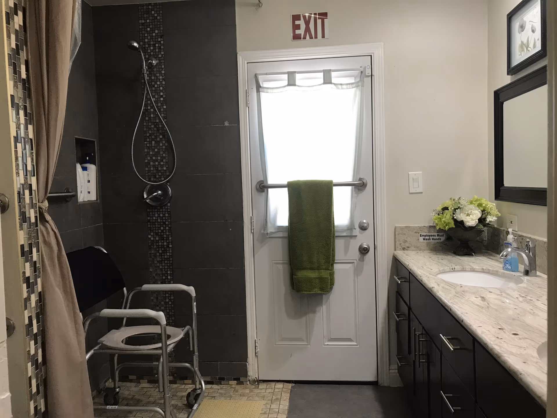 Accessible bathroom with a roll-in shower and shower chair, a sink vanity, and a door with a green towel and EXIT sign.