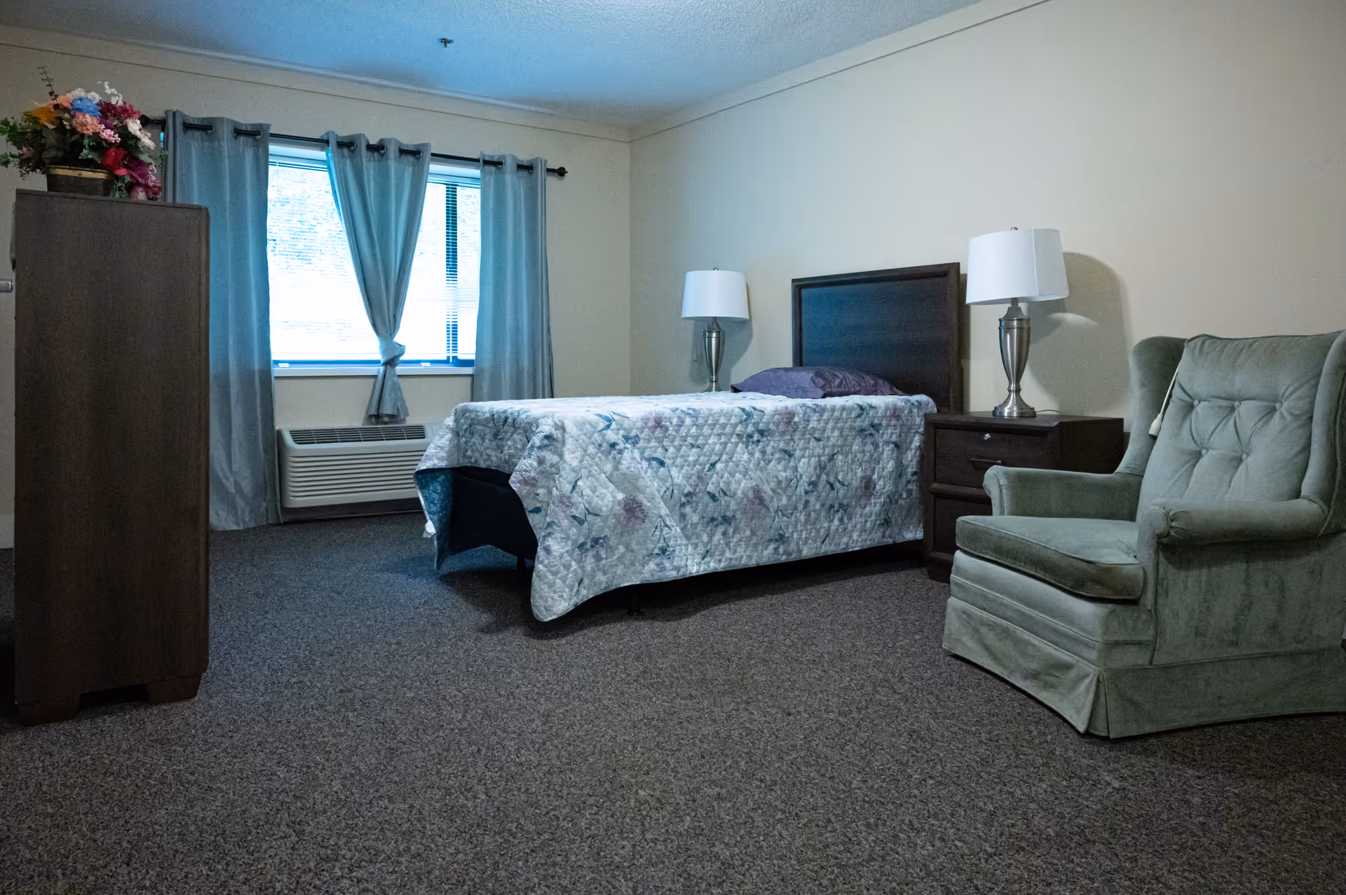A simple bedroom in a senior living facility with a single bed covered by a floral quilt, two bedside tables each with a lamp, a green upholstered armchair, a wooden dresser with a flower arrangement on top, and a window with blue curtains.