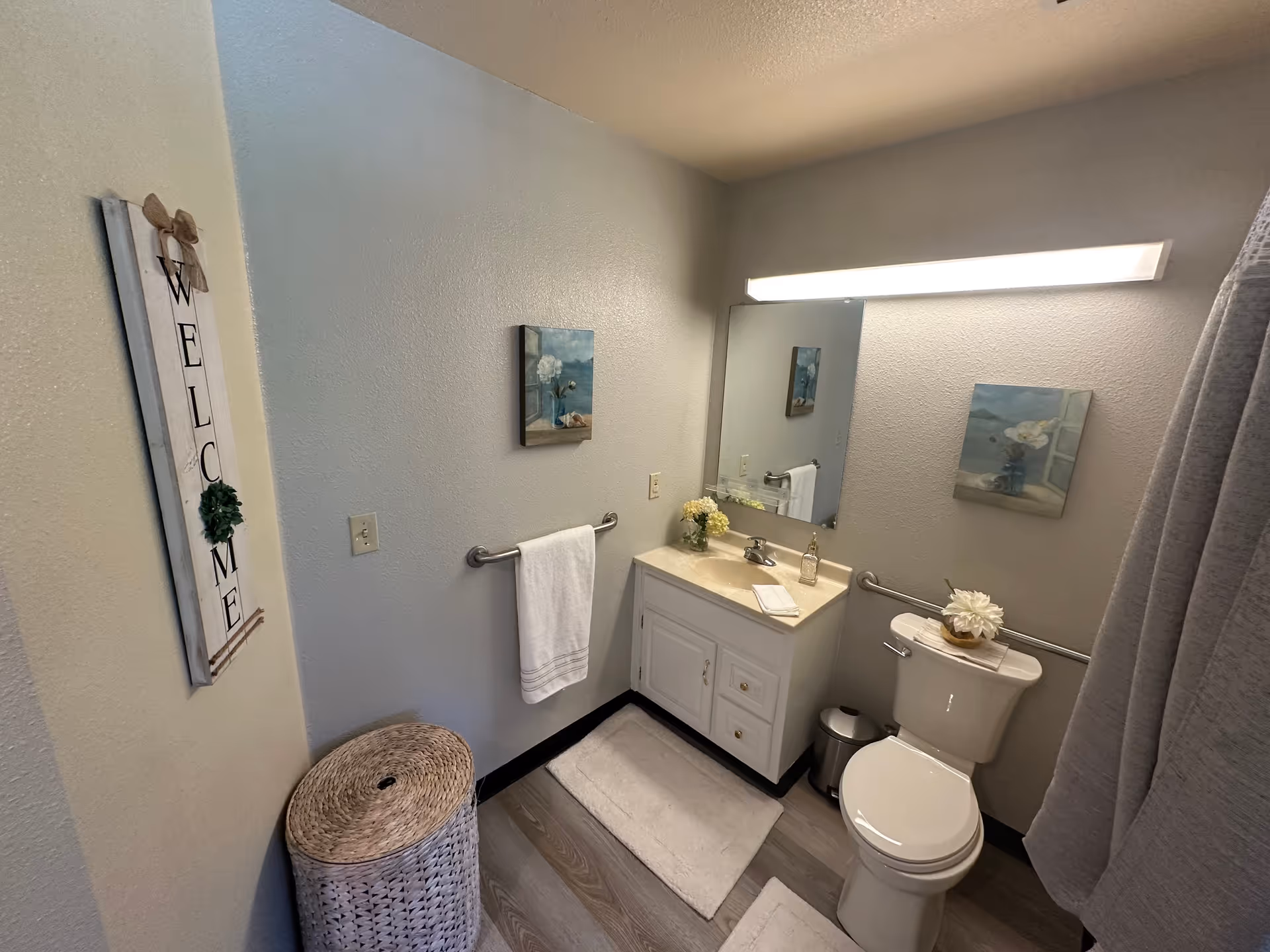 A small, clean bathroom with a white toilet and a white vanity with a sink. Above the vanity is a large mirror with a horizontal light fixture. There is a towel rack with a white towel on the wall, a wicker laundry basket, and decorative wall art including a vertical 'WELCOME' sign and a painting of flowers. The floor has wood-like vinyl planks and there are white bath mats on the floor.