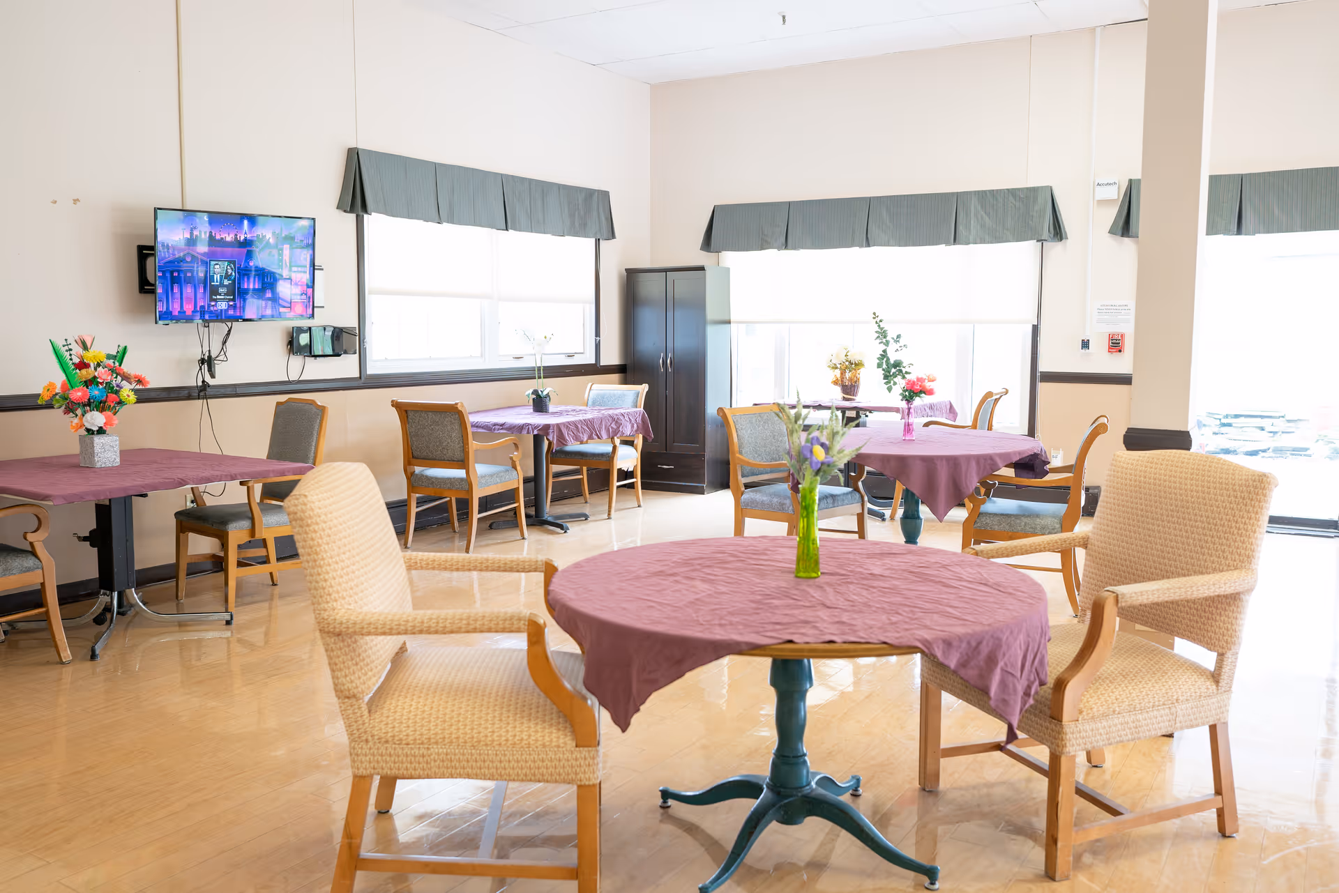 A bright dining room with several tables covered in purple tablecloths, each adorned with a small vase of flowers. There are wooden chairs with cushions around the tables. A flat-screen TV is mounted on the wall, and large windows with green valances allow natural light to fill the room.