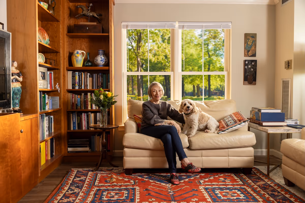 An elderly woman sitting on a beige leather sofa in a cozy living room with a small white dog beside her. Behind them is a large window showing green trees outside. The room features wooden bookshelves filled with books and decorative items, a small round table with a vase of flowers, and a colorful patterned rug on the floor.