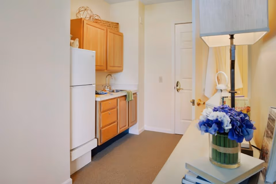 A small kitchen area with wooden cabinets, a white refrigerator, a sink with a green towel hanging on the cabinet handle, and a countertop. In the foreground, there is a table with a lamp, a vase of blue and white flowers, and some books. A closed white door is visible at the end of the kitchen.