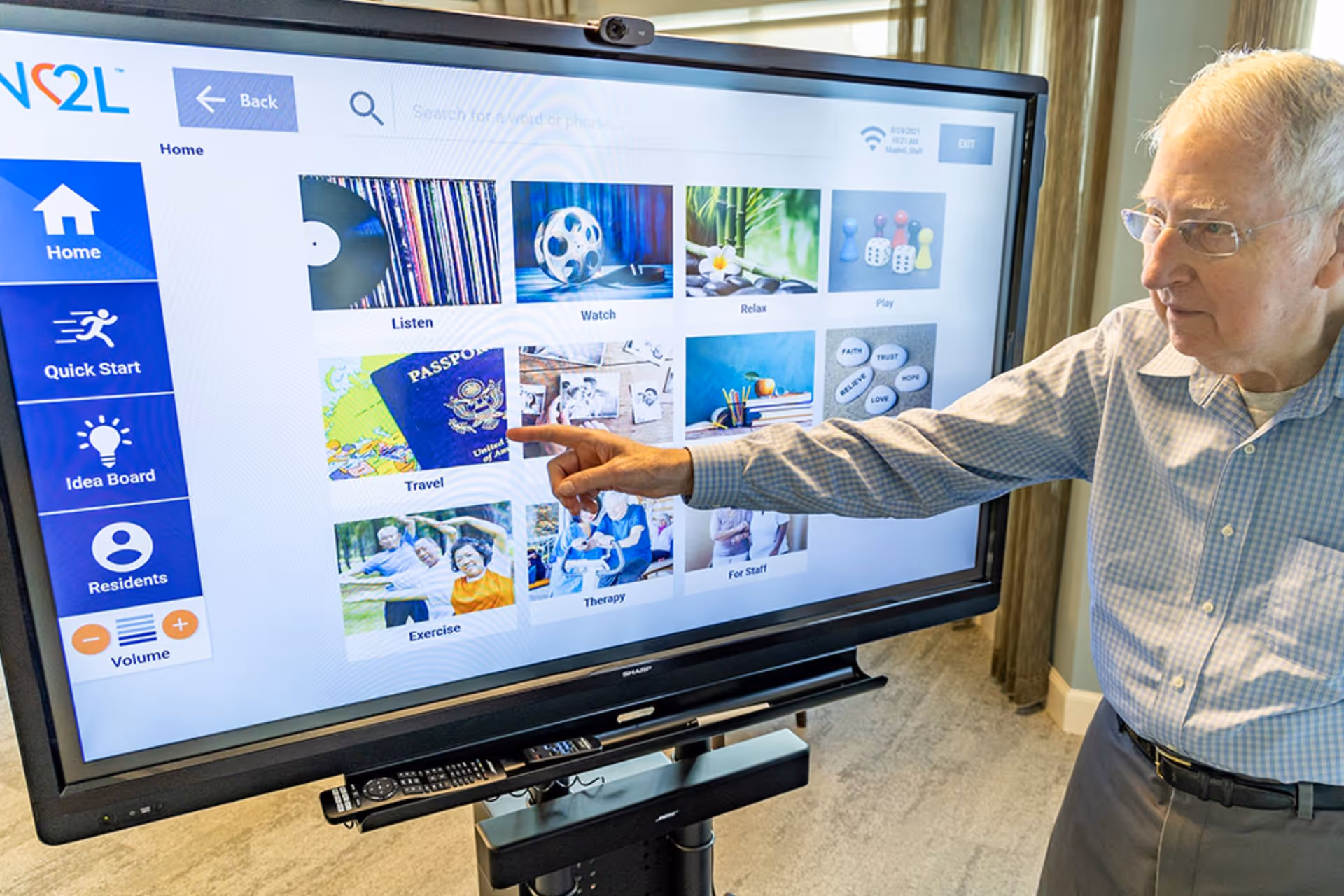 An elderly man wearing glasses and a checkered shirt is interacting with a large touchscreen display showing various categories such as Listen, Watch, Relax, Play, Travel, Exercise, Therapy, and For Staff in a well-lit room with curtains in the background.