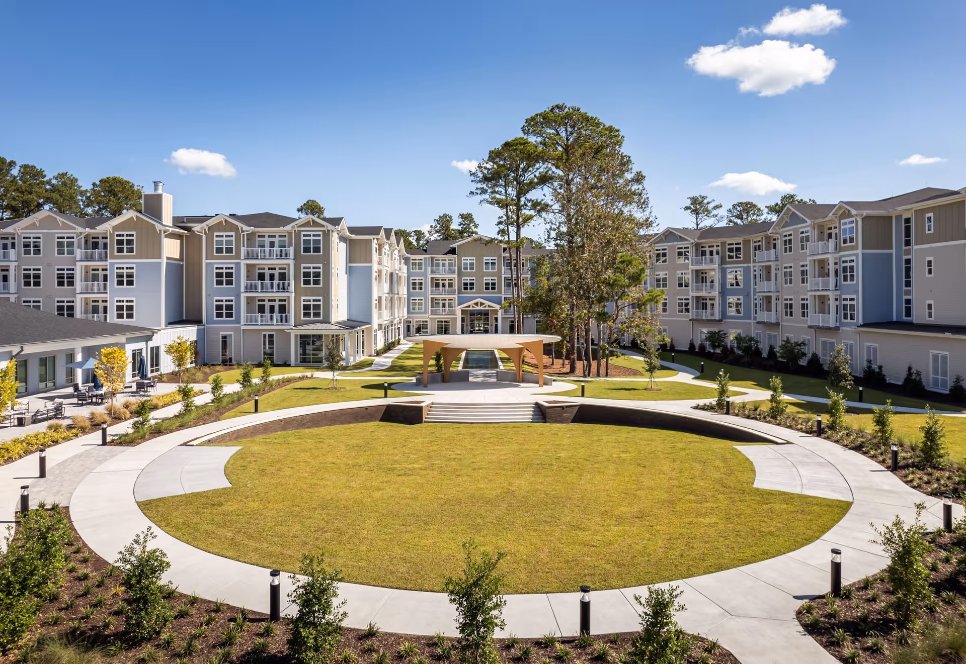 A large, circular grassy courtyard surrounded by a paved walkway and landscaped with small bushes and trees. The courtyard is part of a senior living community with multi-story residential buildings in the background under a clear blue sky with a few clouds.