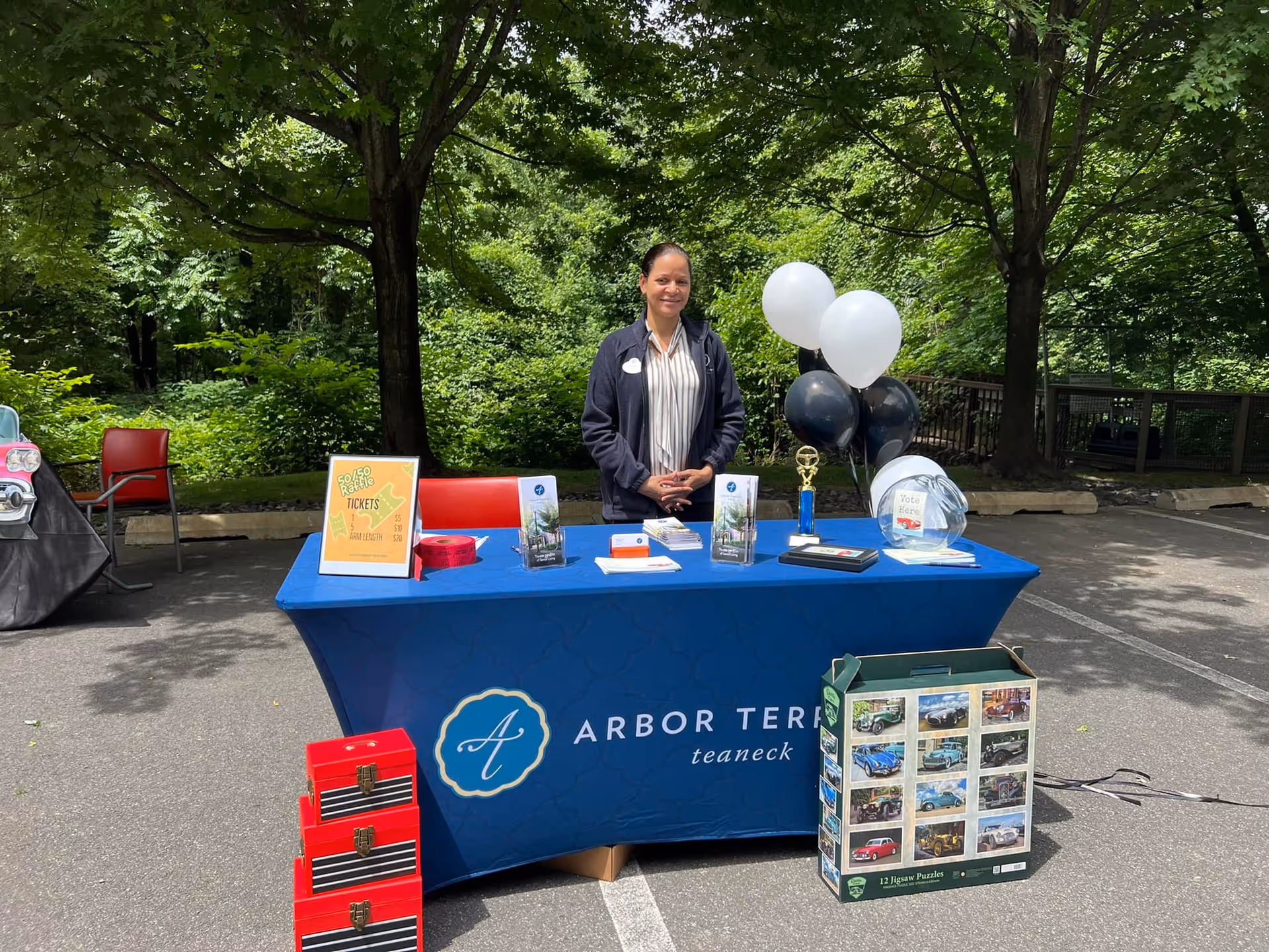 A woman stands behind a blue table covered with promotional materials and balloons outdoors. The tablecloth has the Arbor Terrace Teaneck logo. There are trees and greenery in the background, and the setting appears to be a parking lot or paved area.