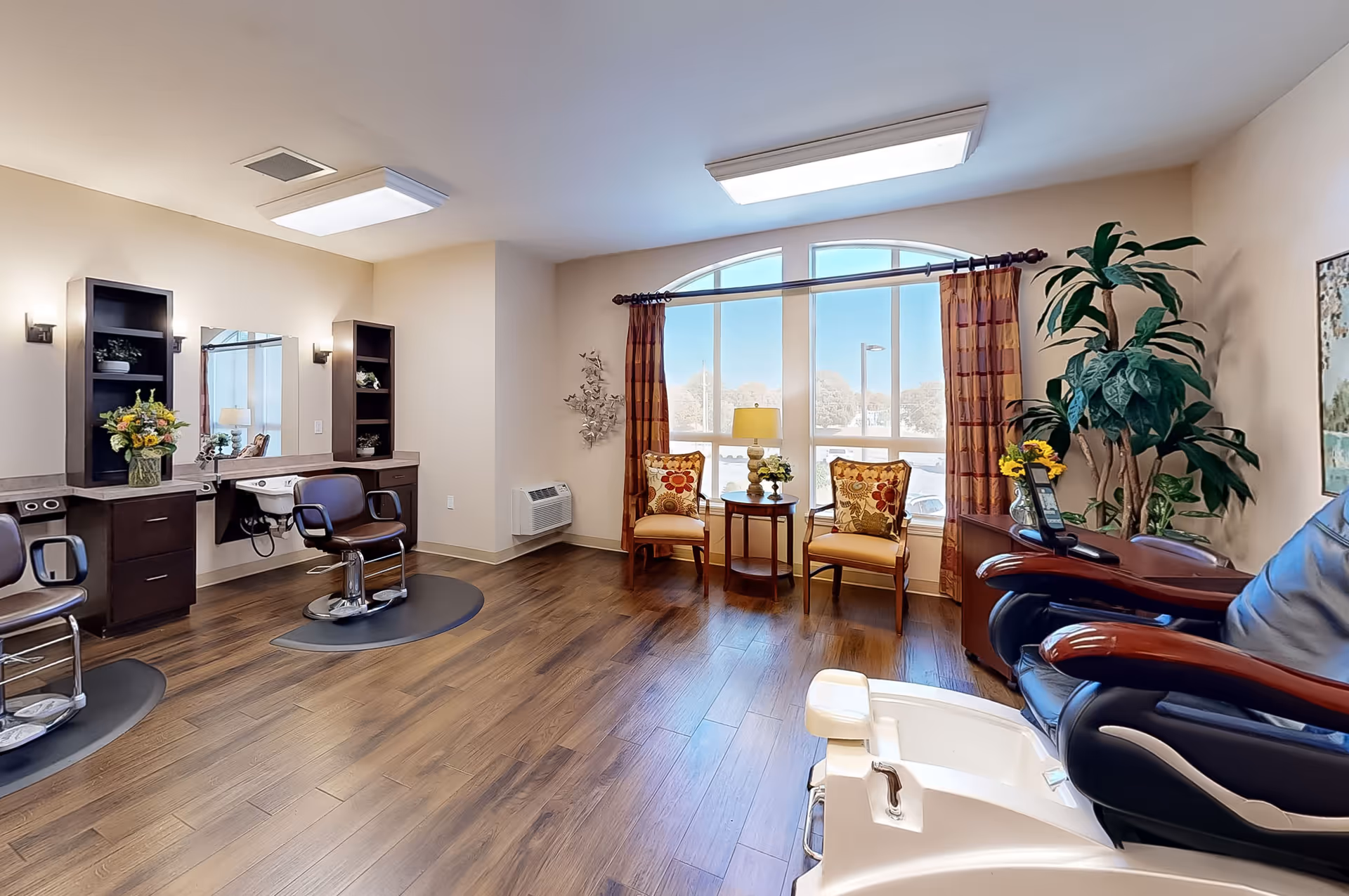 Interior view of a salon area in an assisted living facility featuring two salon chairs with mirrors and shelves, a pedicure chair in the foreground, two cushioned chairs with floral pillows near a large window with curtains, a small round table with a lamp and flowers, and a large potted plant in the corner.