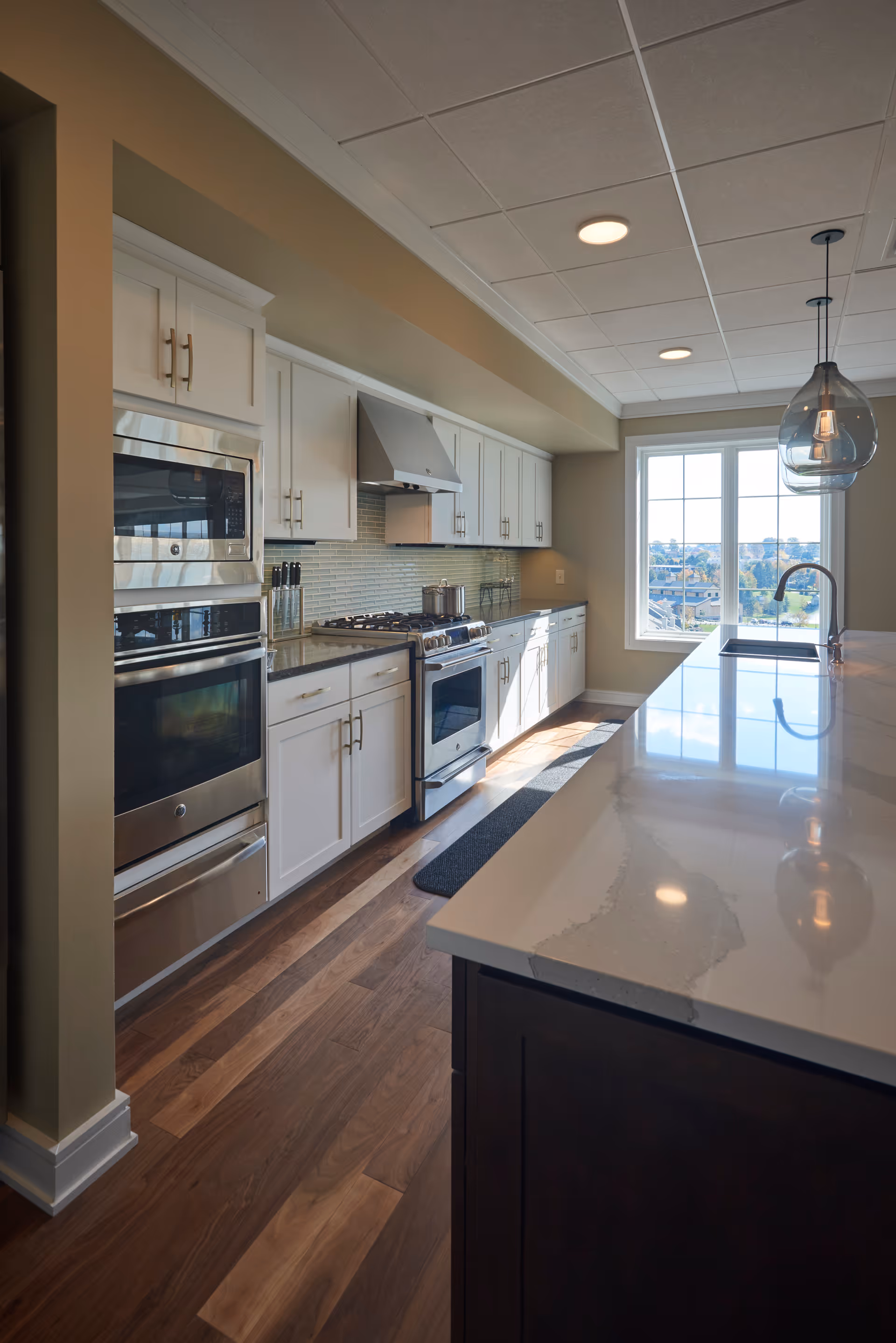 Modern kitchen with white cabinets, stainless steel appliances including a microwave and oven, a gas stove with a range hood, and a large island with a marble countertop and a sink. The kitchen has wooden flooring and a large window letting in natural light.